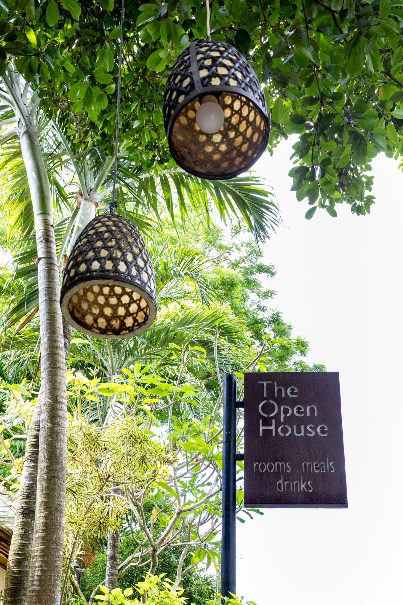 Outdoor scene with leafy green trees, hanging lanterns, and a sign that reads "The Open House" with listings for rooms, meals, drinks.