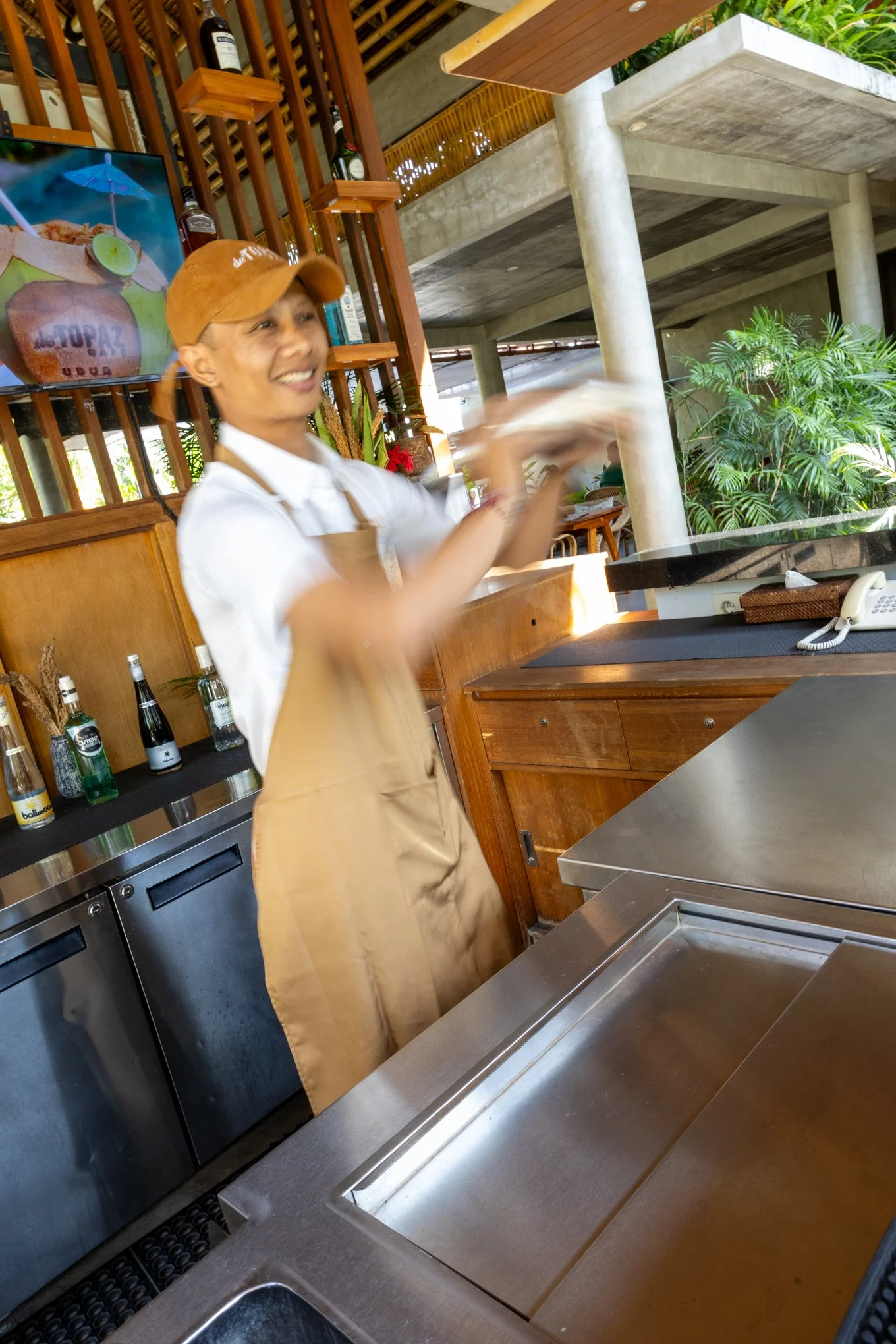A smiling young woman wearing a brown baseball cap and beige apron standing behind a stainless steel kitchen counter with a sink, in a restaurant or cafe with wooden shelves holding bottles, and green plants in the background.