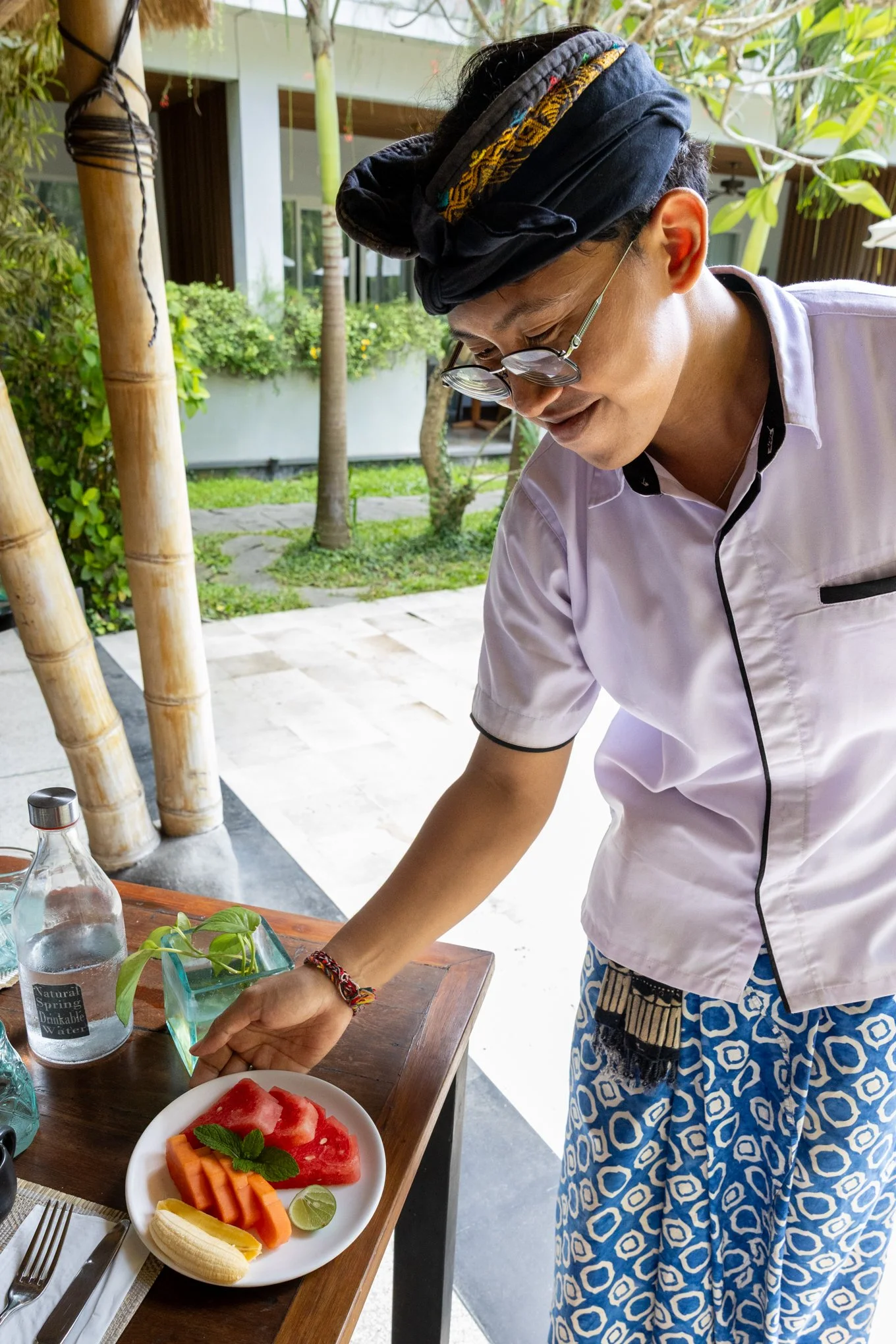 A person wearing glasses and a black turban, dressed in a white shirt and blue patterned sarong, is preparing food outdoors on a wooden table with tropical plants and bamboo in the background. There are sliced fruits on a white plate and bottles of w