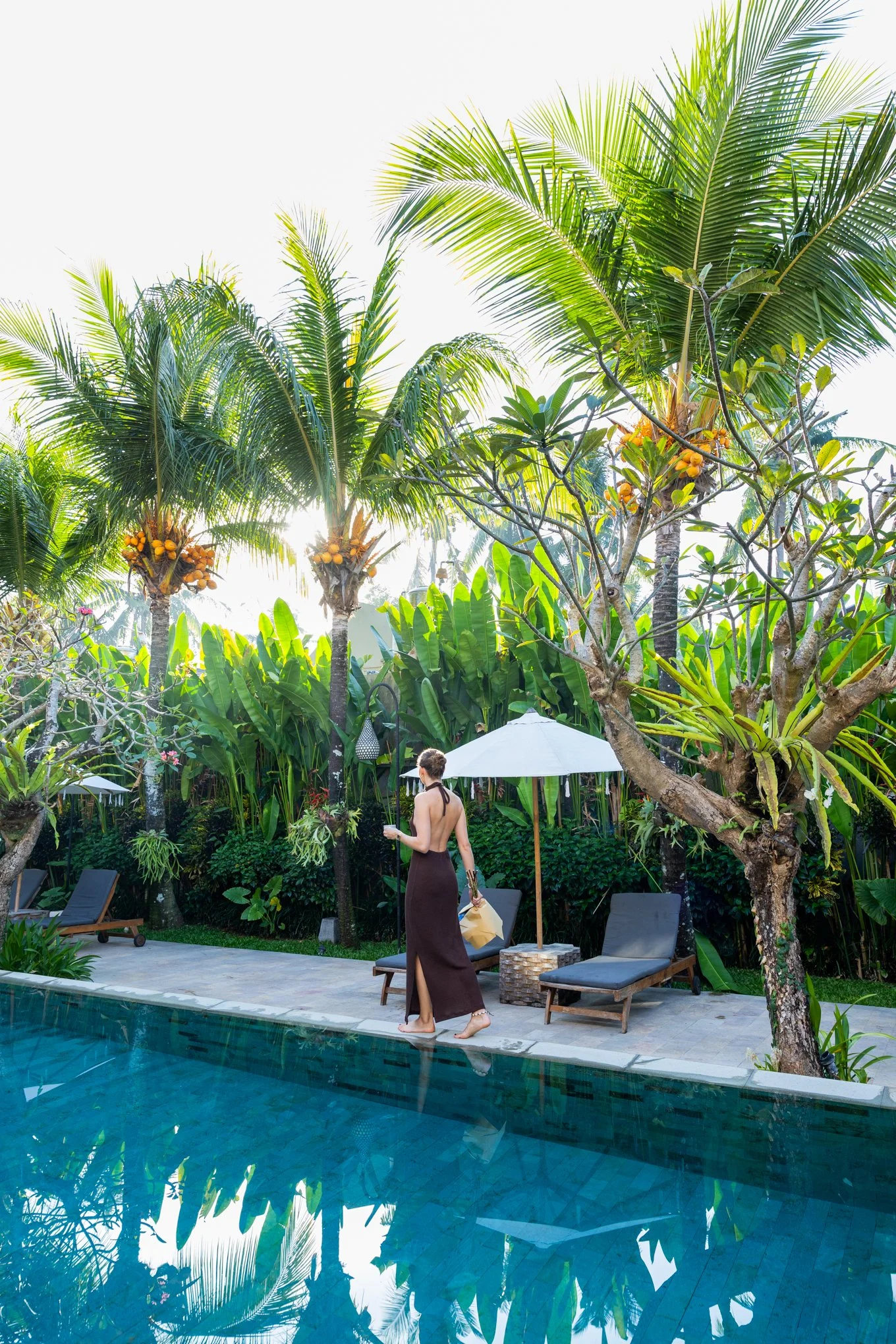 A woman wearing a long brown skirt and black bikini top walking barefoot along the edge of a swimming pool. The scene is set in a lush tropical garden with palm trees, other green plants, and white umbrellas providing shade. The pool water reflects the surrounding plants and sky, creating a peaceful atmosphere.