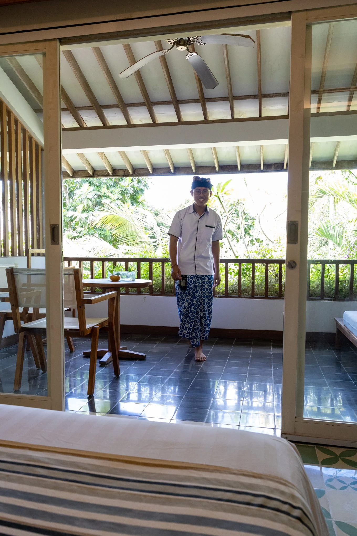 A man standing barefoot on a balcony with green foliage in the background. He is dressed in traditional attire with a white shirt and patterned sarong, wearing glasses and smiling.