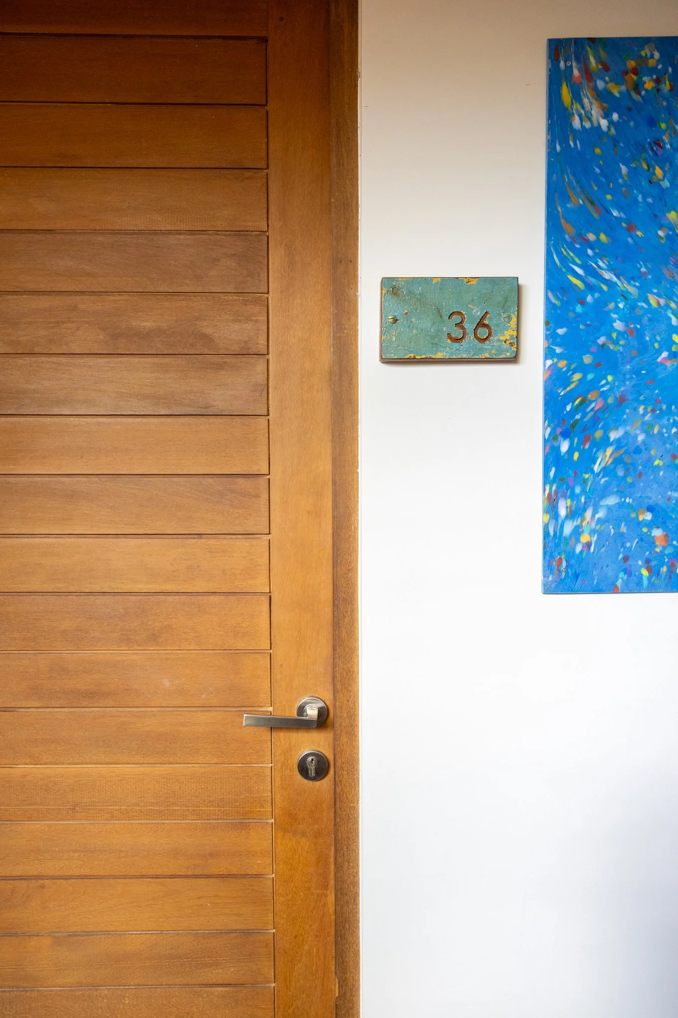 A wooden door with a silver handle and a keyhole, next to a white wall decorated with a blue abstract painting and a small weathered wooden sign with the number 36.
