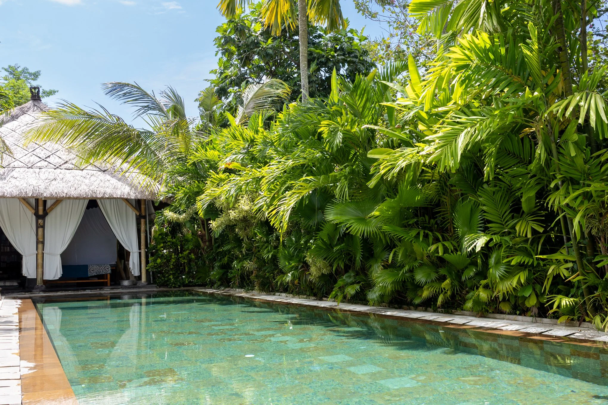 A tropical scene featuring a swimming pool with crystal-clear water, lush green palm trees, and tropical plants surrounding a thatched-roof hut with white curtains.