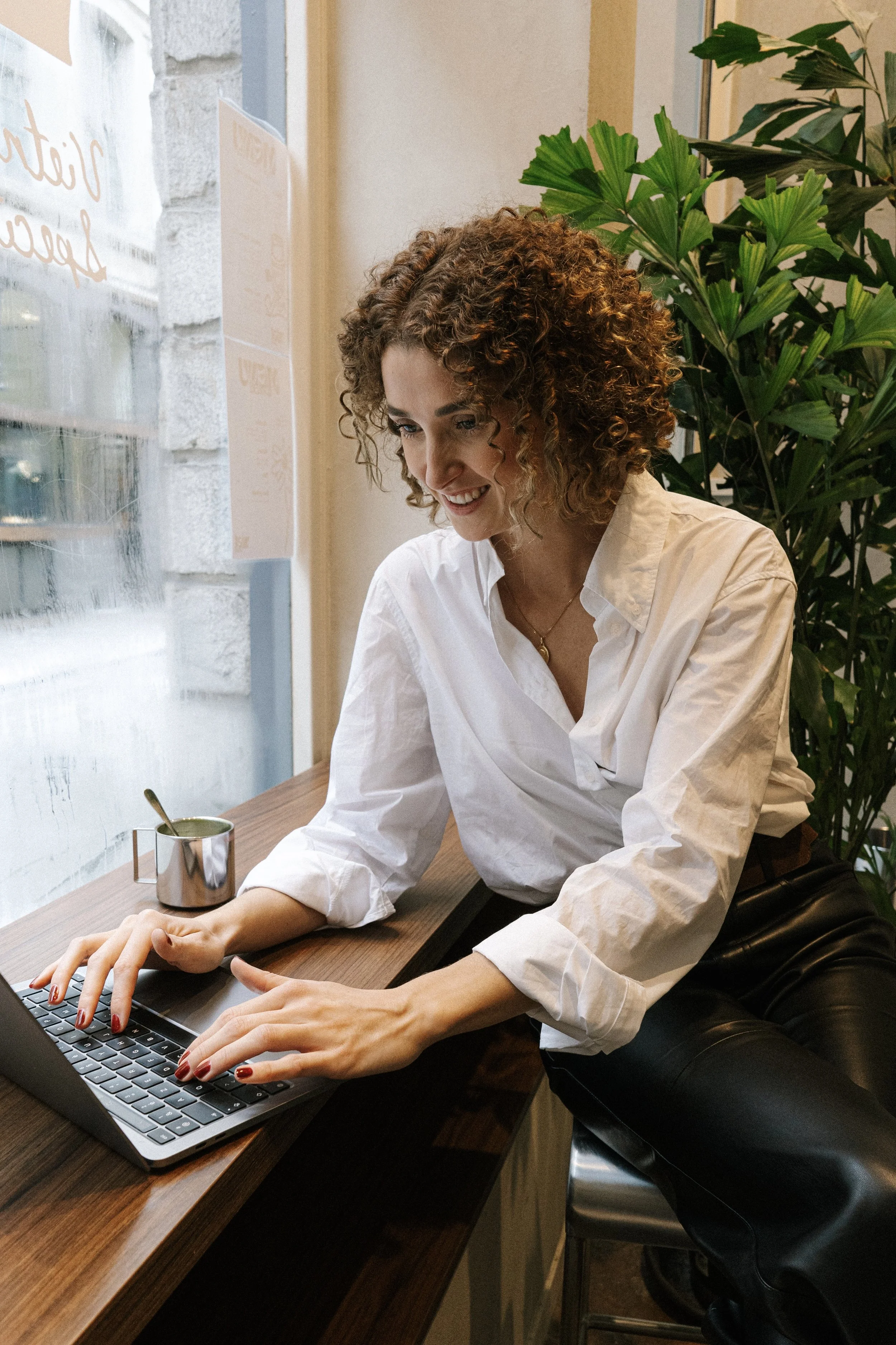 A woman with curly brown hair wearing a white blouse is sitting at a wooden table, using a laptop. She is smiling and appears to be working or browsing. Beside her is a metallic cup with a spoon. Behind her, there are large green plants and a window with light coming through.