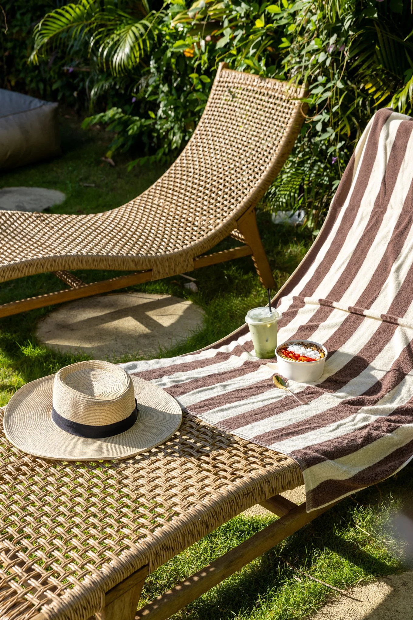 A wicker lounge chair and a striped towel on a lounge chair, with a straw hat, a drink, and a bowl of snacks on the towel, set outdoors on a sunny day with lush green plants in the background.