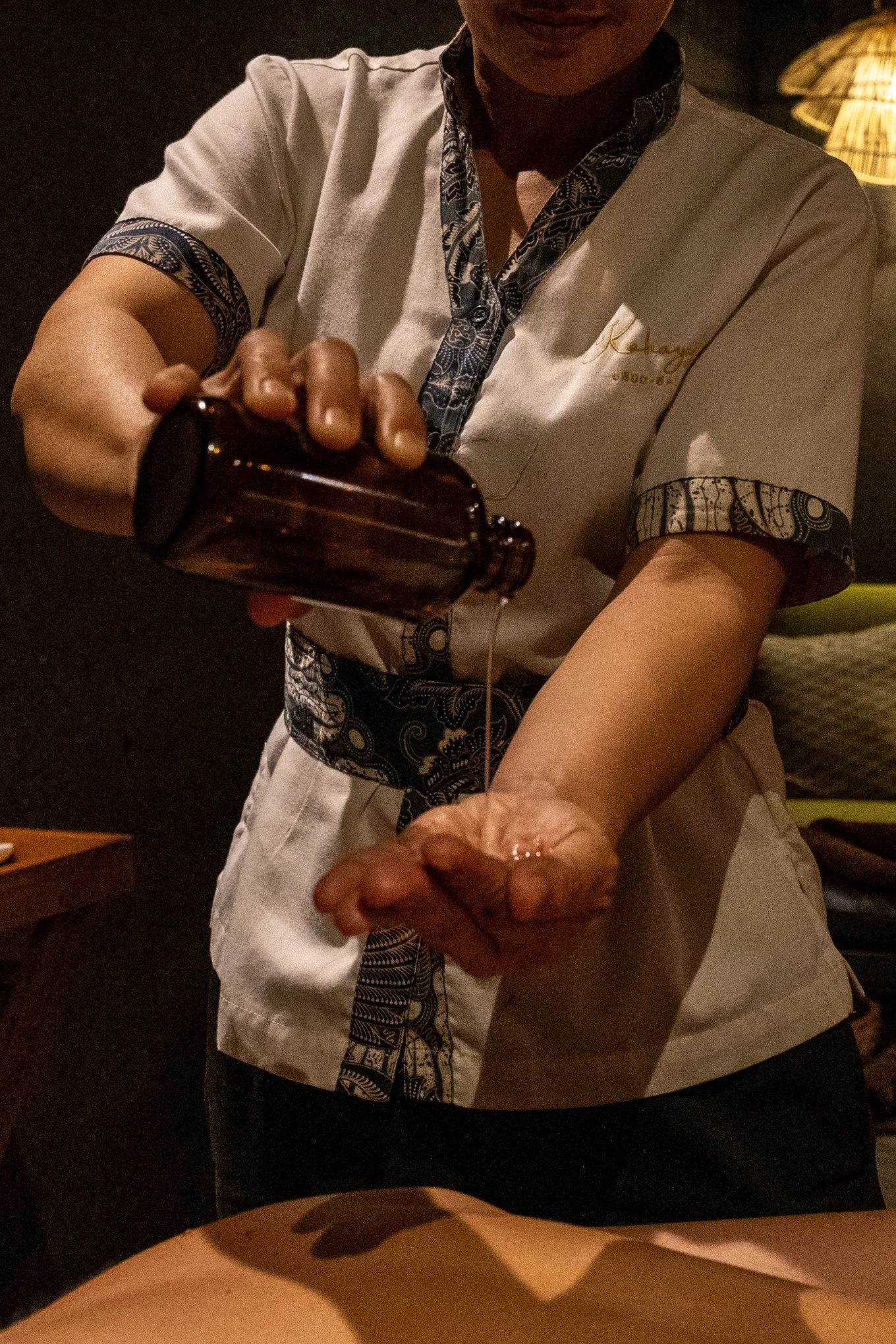 A person in a white uniform with black and white patterned trim, holding a small dark bottle with a dropper and administering a liquid to someone else's arm.