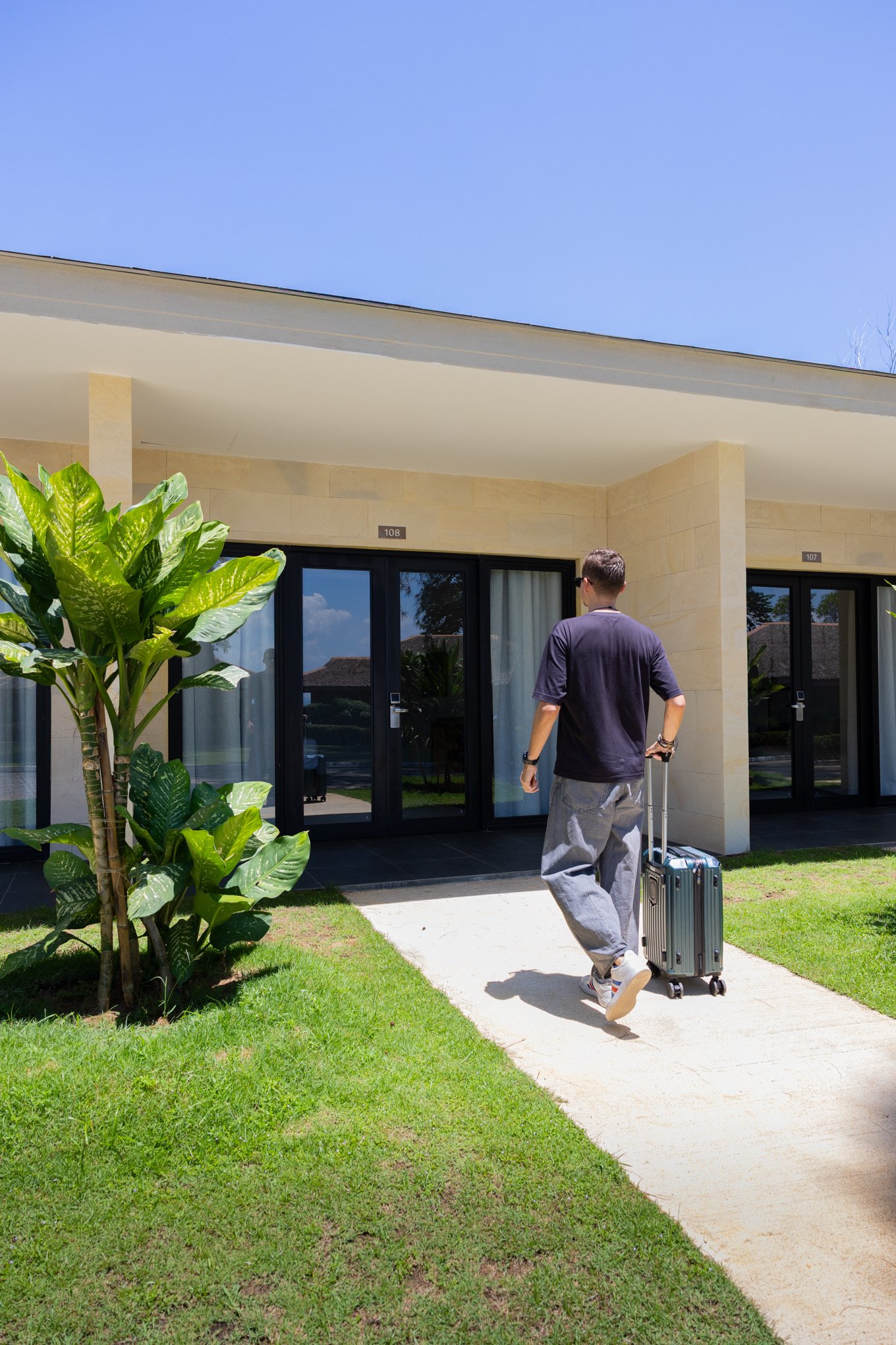 A man walking towards a modern building with glass doors, carrying a rolling suitcase, on a sunny day with a well-maintained lawn and large green plants nearby.