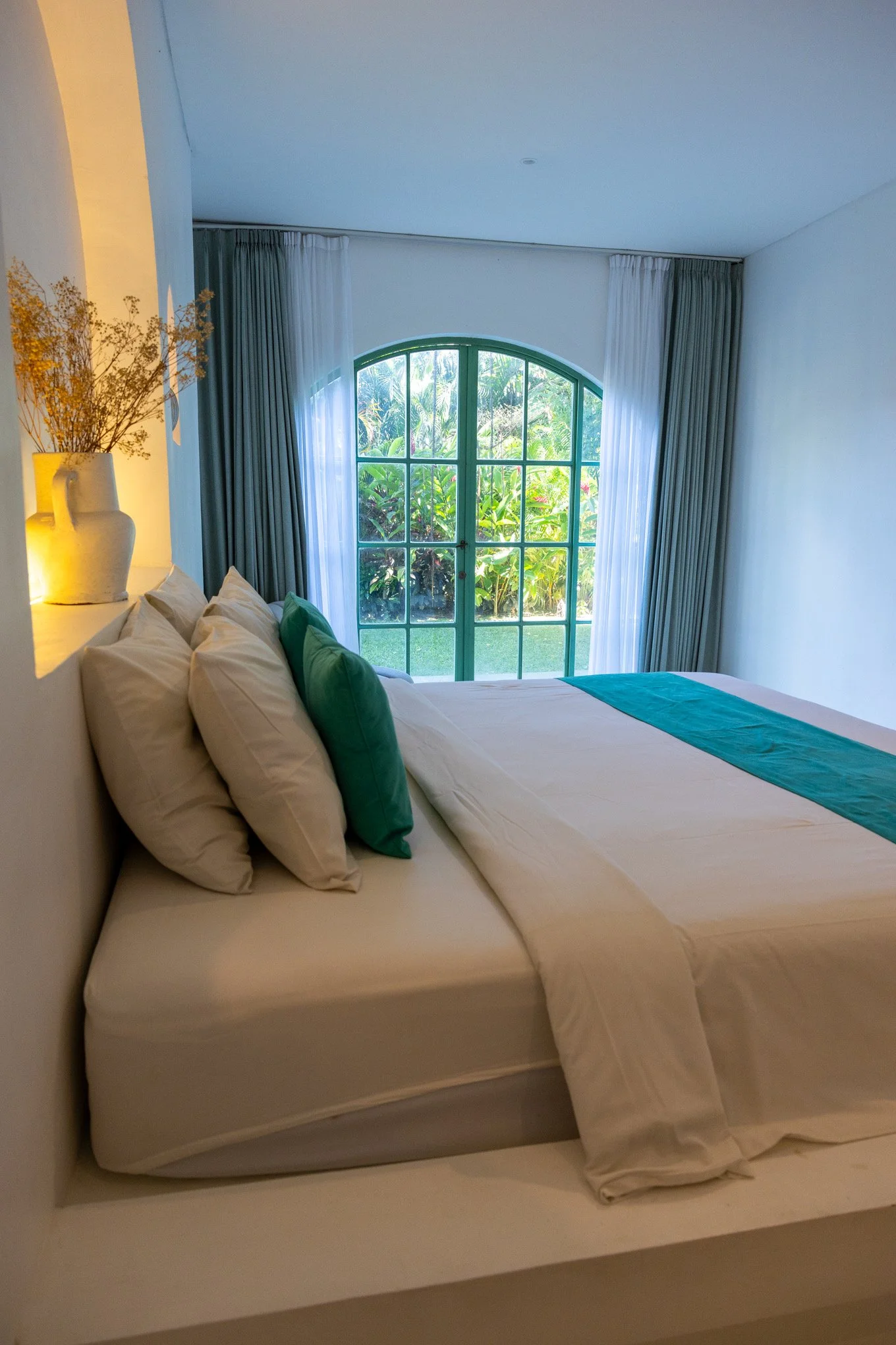 A neatly made bed with white sheets, white and green pillows, located in front of a large window with white and green curtains, overlooking a garden with green plants.