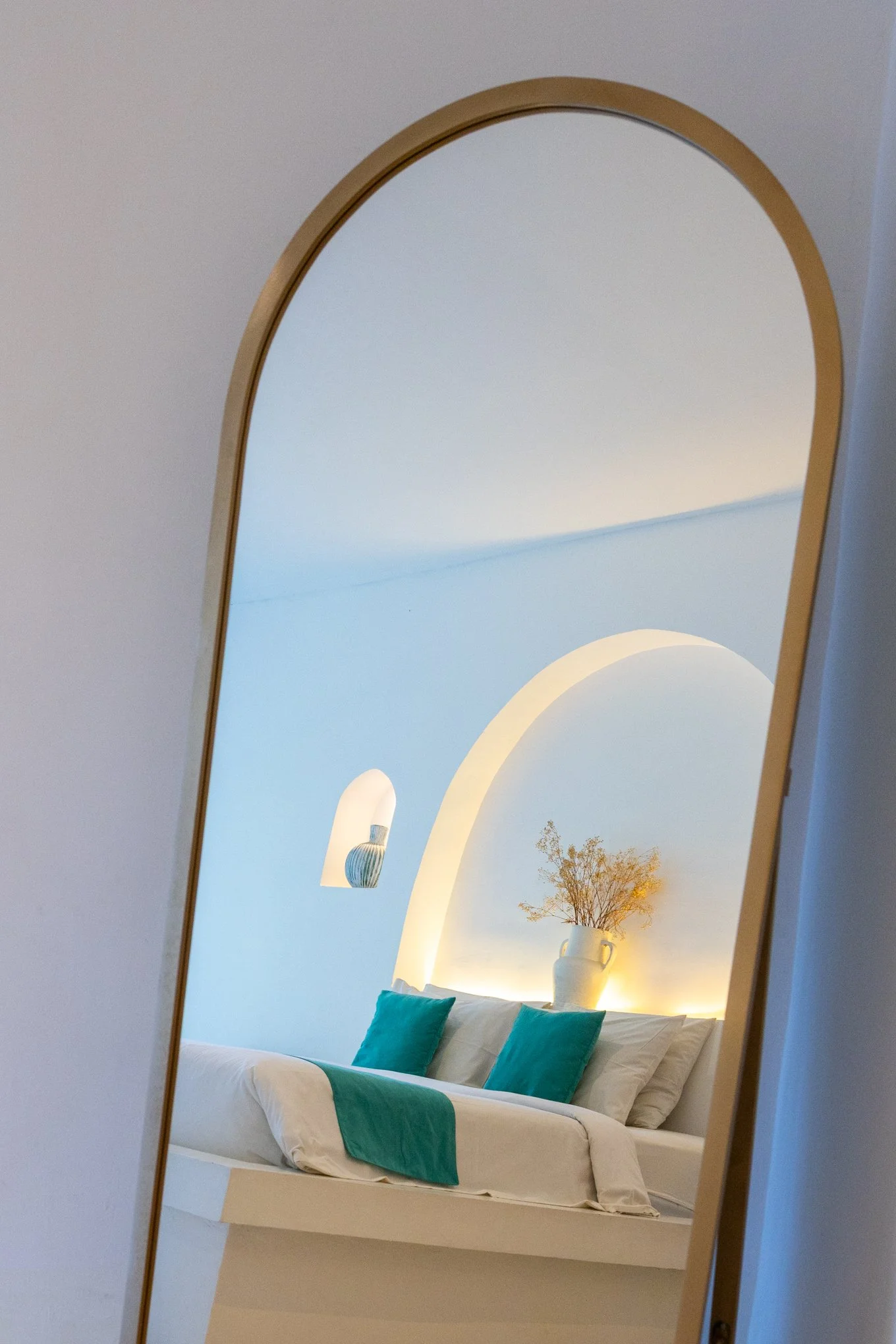 A bedroom with a mirror showing a white bed with teal pillows, a softly lit wall with an arched niche containing a ceramic vase and dried flowers, and a small plant in a pot on a shelf.