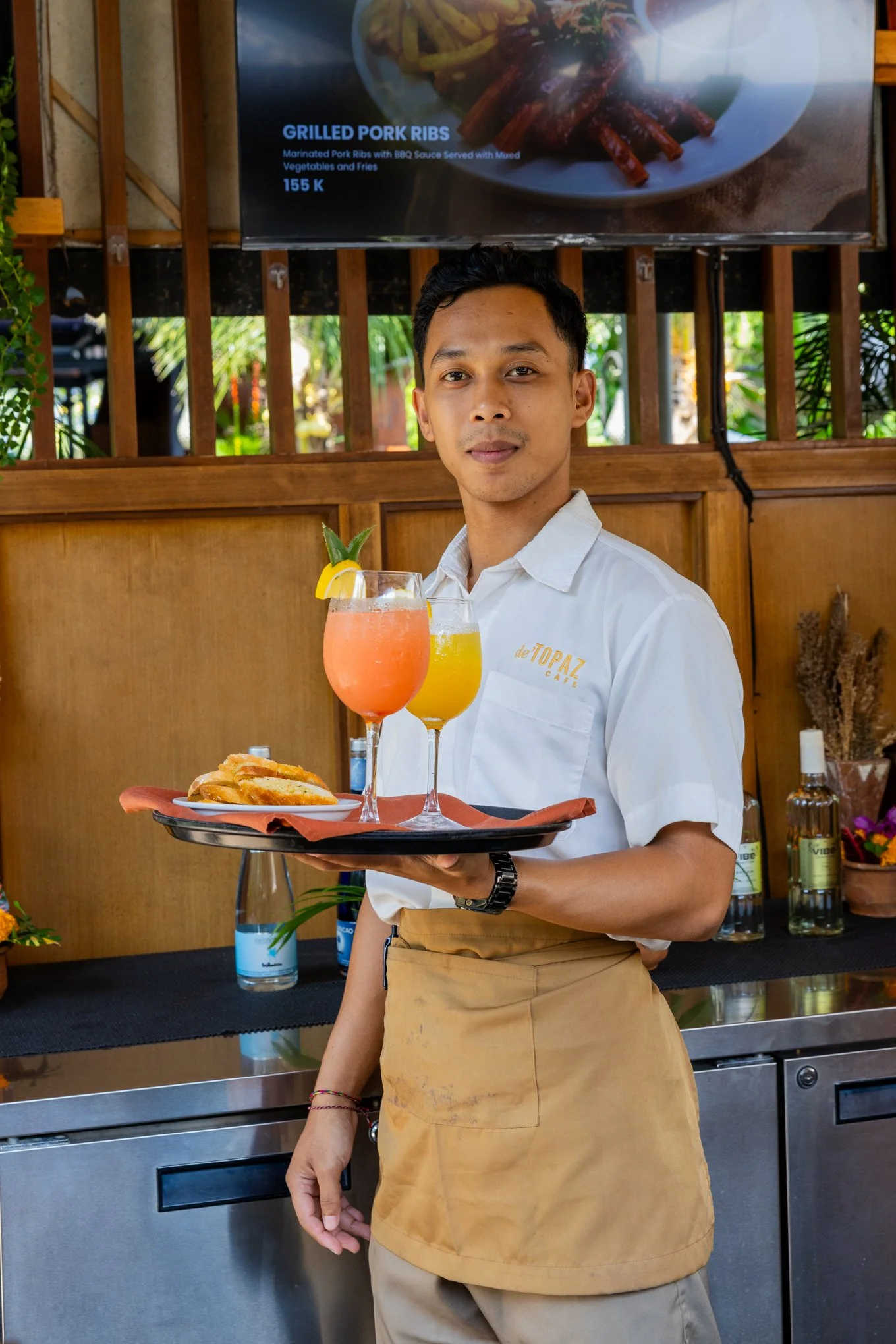 A smiling waiter in a white shirt and tan apron holding a tray with grilled pork ribs, a slice of lemon-lime garnish, and a pink cocktail in a glass, in a restaurant setting with a wooden wall and a menu screen displaying grilled pork ribs.