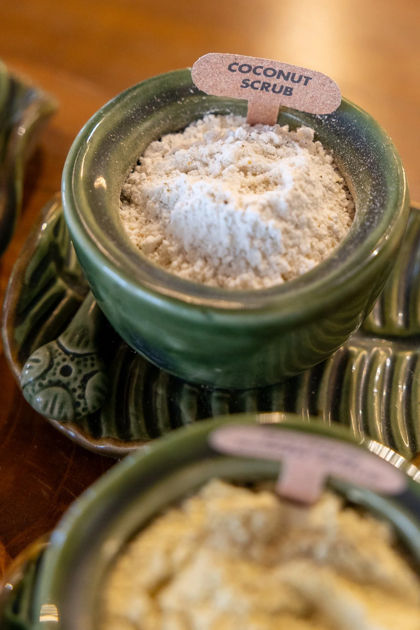 Close-up of a small green ceramic jar filled with coconut scrub labeled with a small tag.