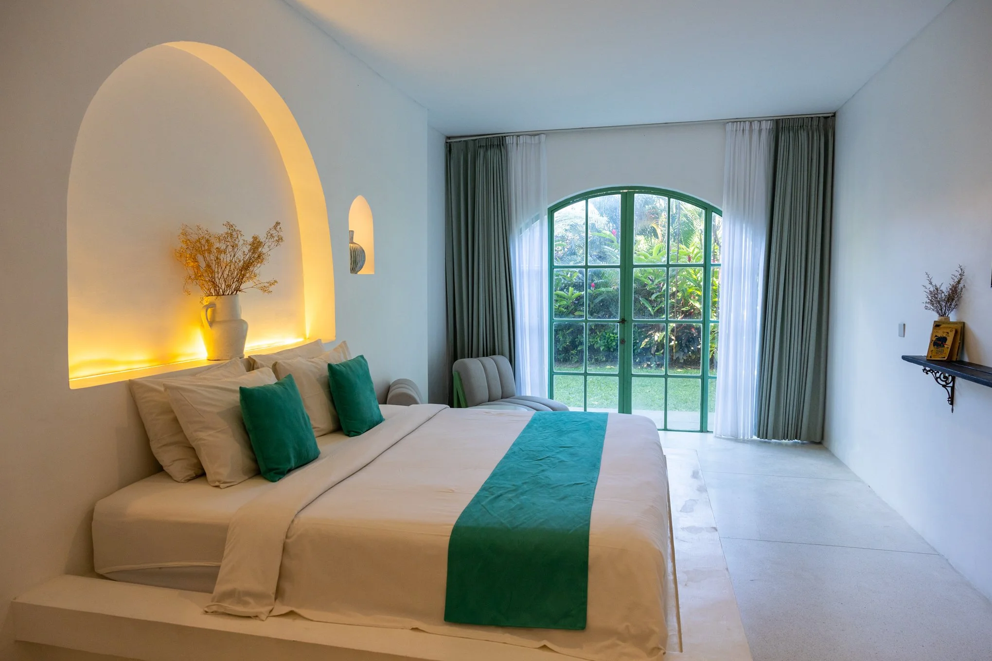 A bright bedroom with a large bed adorned with white and green pillows, a teal runner, and a decorative throw pillow, by a window with green framing and view of lush greenery outside, with a white curtain, and a shelf with a small decorative item.