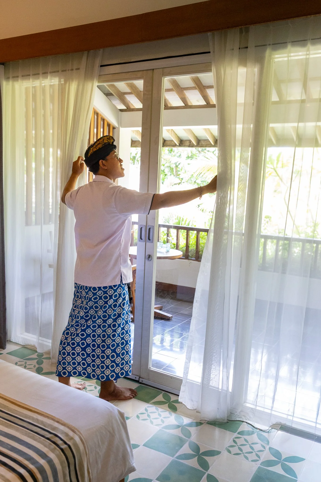 A woman dressed in a pink shirt and a blue patterned skirt, wearing glasses and a headscarf, is standing barefoot at a sliding glass door, pulling back sheer curtains to look outside. The room has tiled floors and the balcony outside has wooden beams