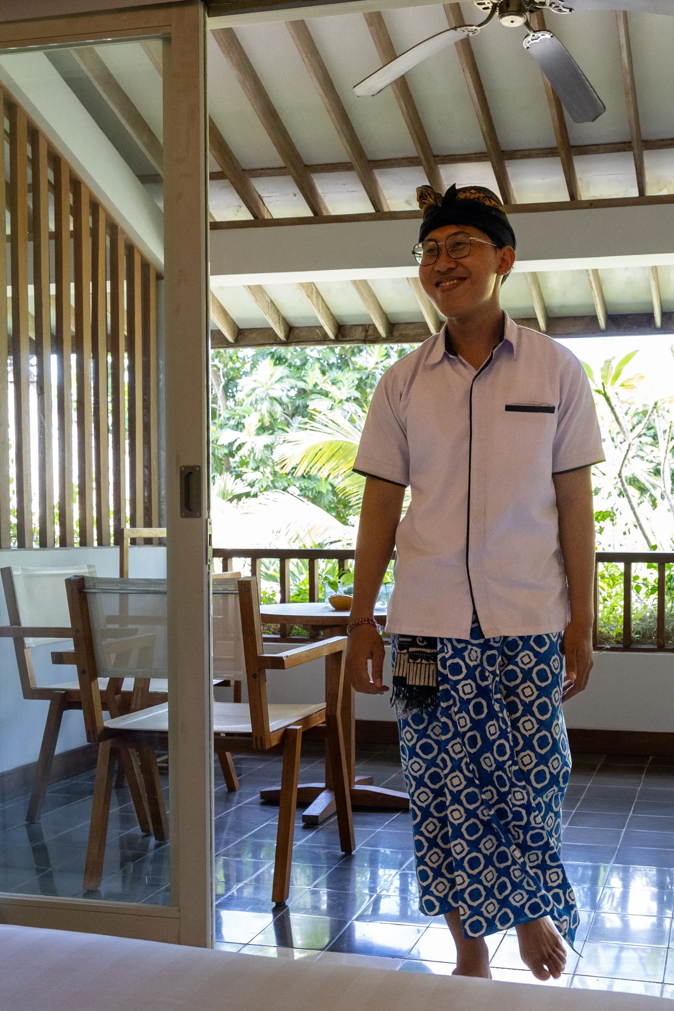 A smiling man with glasses and a black head wrap, standing indoors near a sliding glass door, wearing a light pink short-sleeved shirt and patterned blue and white sarong with a tropical outdoor scene visible through the window.