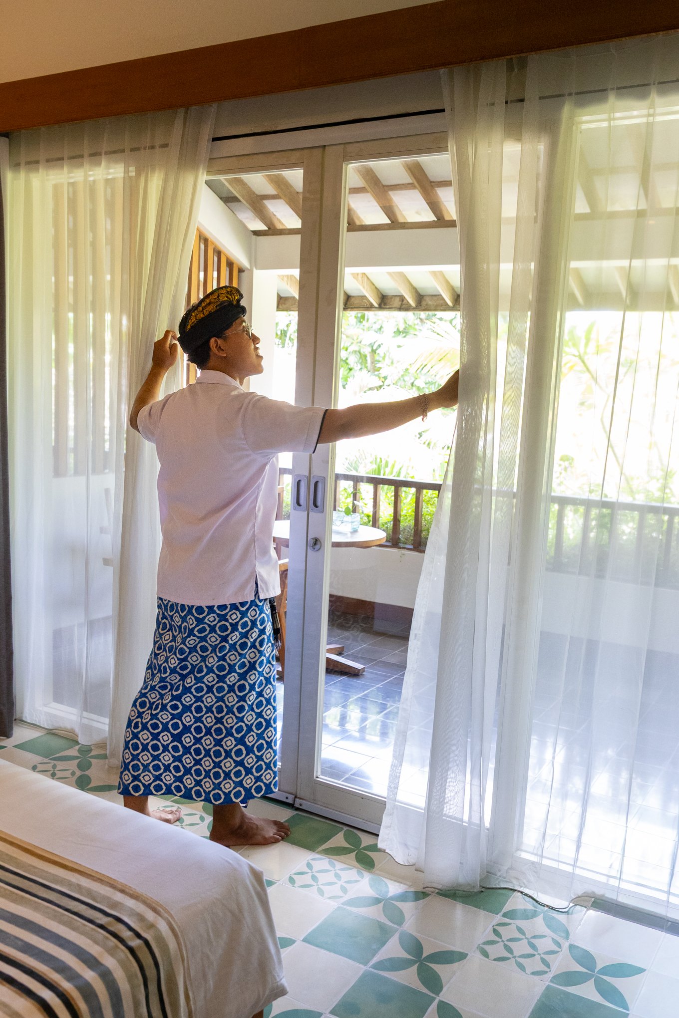 A woman standing inside a room, opening sheer curtains and looking outside at a balcony with a rustic roof and greenery beyond.