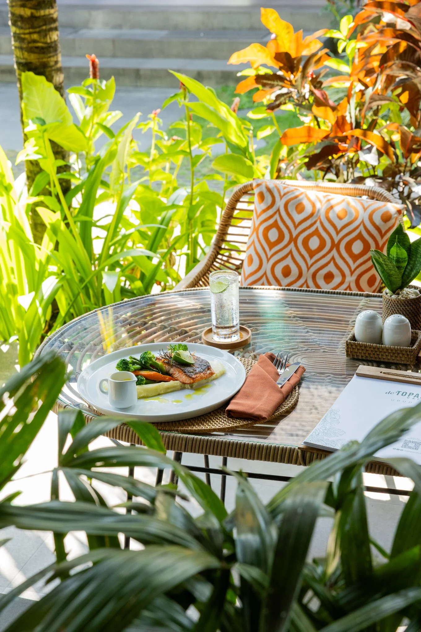 A tropical outdoor dining setup with a glass of water with lime, a plate of fish with vegetables, a woven table, a patterned cushion on a wicker chair, and various green plants surrounding the area.