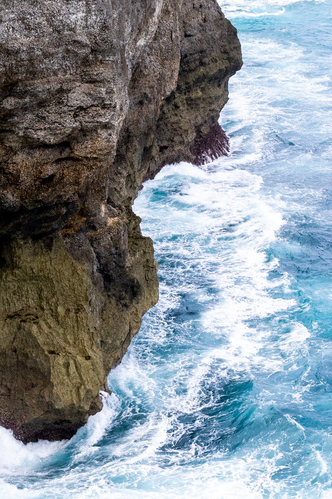 Close-up of a rocky cliff face with rough texture, meeting turbulent blue ocean water with white foam at the base.