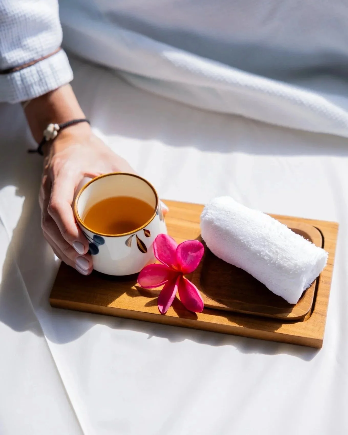 A hand holds a cup of tea on a wooden tray with a white towel, pink flower, and white napkin, with a white cloth background.