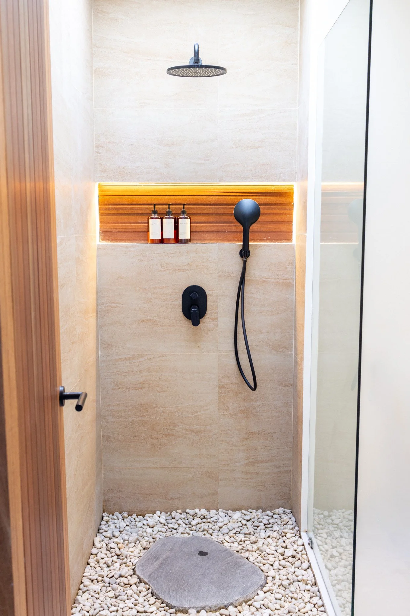 Modern outdoor shower with a rainfall showerhead, handheld shower, three shampoo bottles, and a stepping stone in a pebble-filled shower area with glass walls and wood accents.