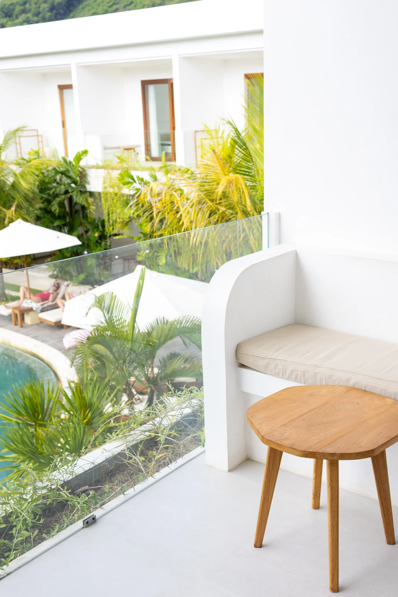 A white balcony with a round wooden table and a built-in white cushioned seat. Behind the balcony, there are tropical plants and a view of an outdoor communal pool area with lounge chairs, umbrellas, and greenery.