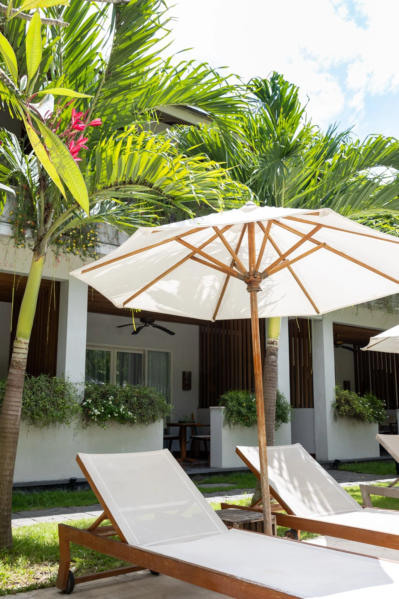 Poolside outdoor area with white lounge chairs and a large white patio umbrella, surrounded by tropical green plants and trees.