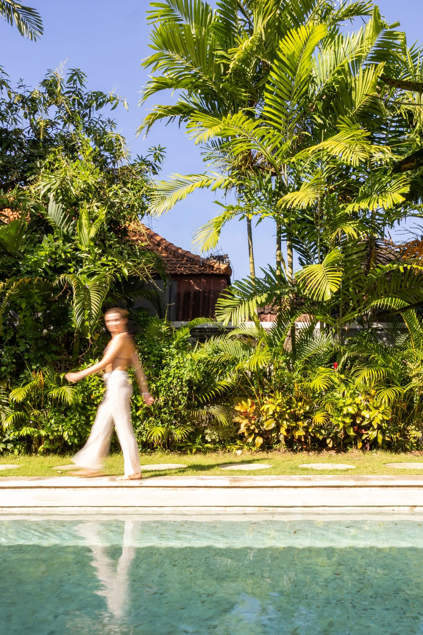 Woman walking along a tiled pathway next to a swimming pool, with lush green tropical plants and palm trees in the background under a clear blue sky.