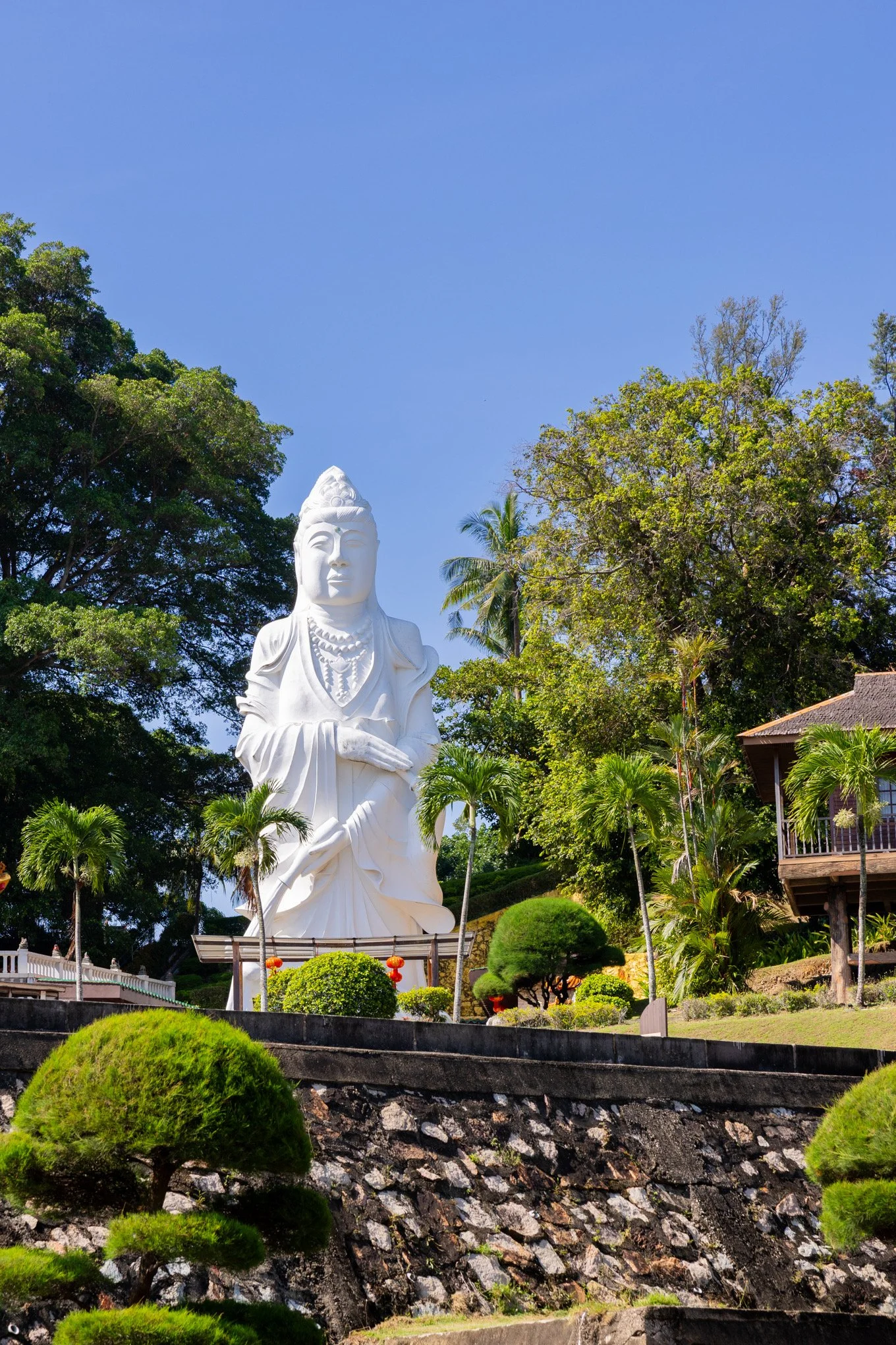 A white statue of a woman with a calm expression, wearing jewelry and a crown, stands amid lush green trees and plants under a clear blue sky.