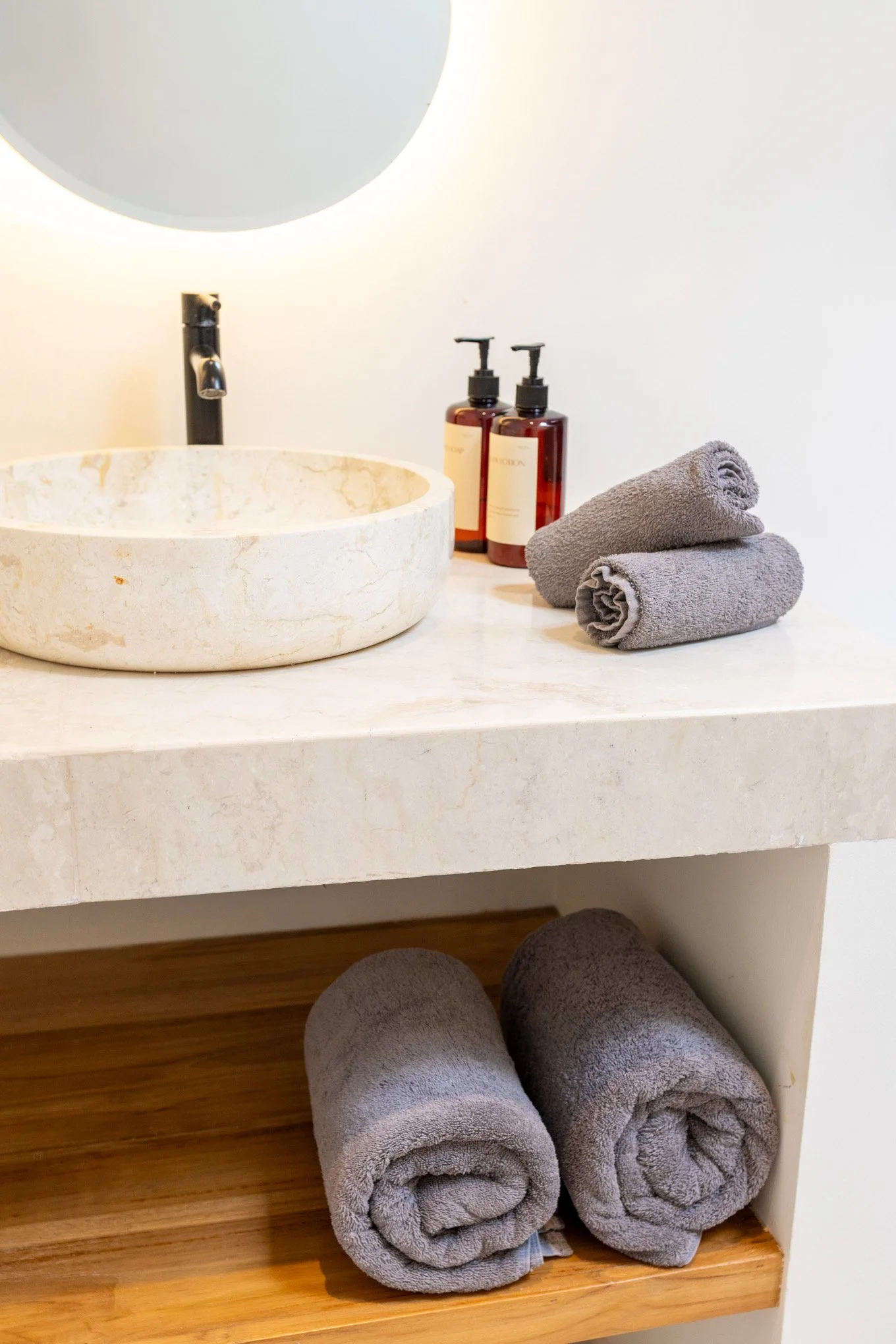 Bathroom countertop with a round marble vessel sink, a black faucet, two amber pump bottles, and three rolled gray towels stacked on the side, with wooden flooring below.