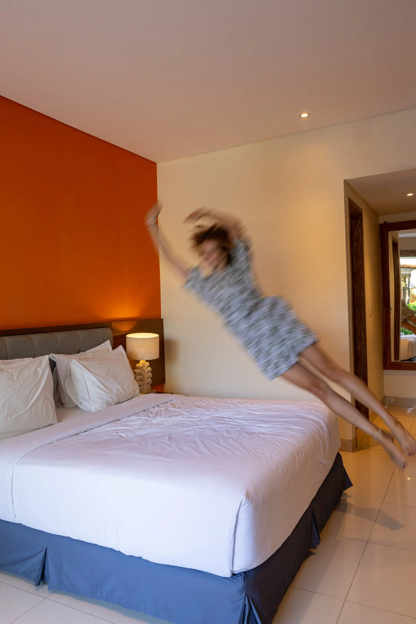 A woman jumping on a bed in a hotel room, wearing pajamas, with a white bed, pillows, and a nightstand with a lamp, in a room with orange and white walls.