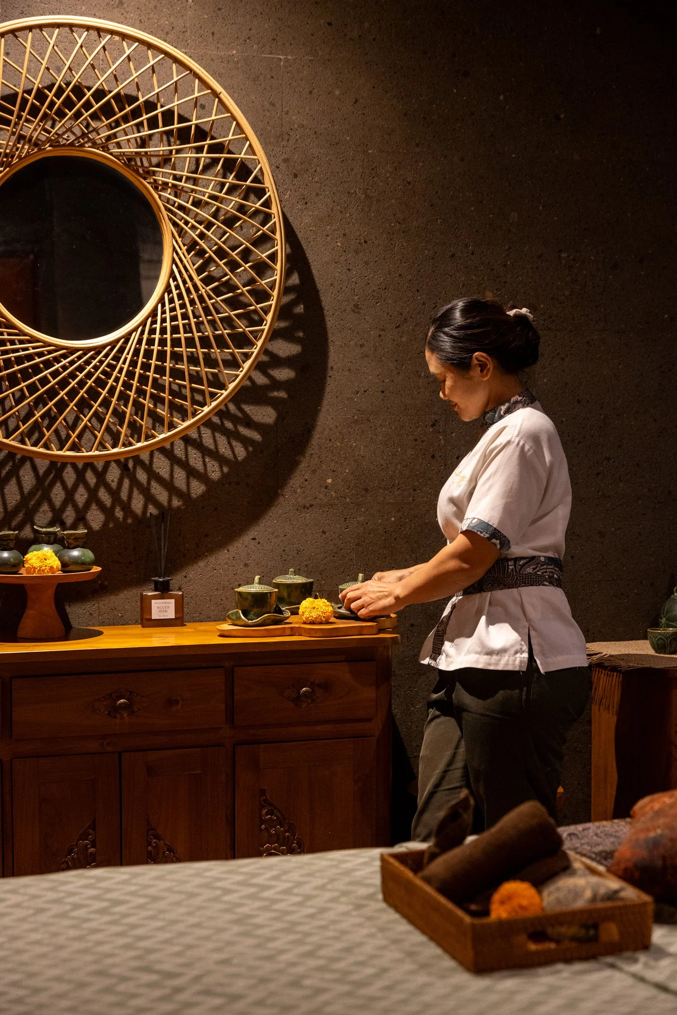 A woman in traditional attire preparing tea or making a traditional craft in a dimly lit room with a textured wall. There are various dishes and decorative items on the wooden furniture.