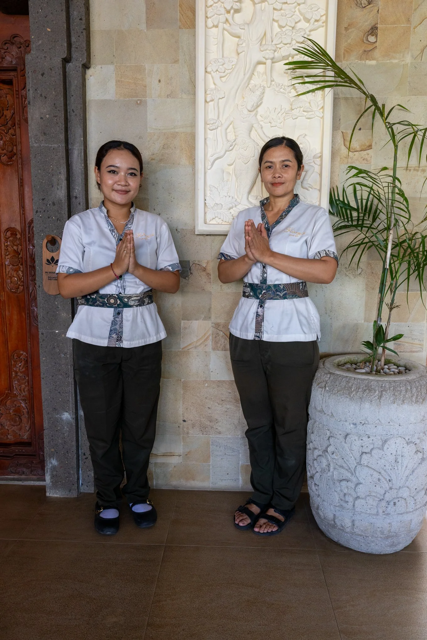 Two women standing side by side in traditional attire, hands pressed together in a prayer gesture, in front of a carved stone relief and a large potted plant.