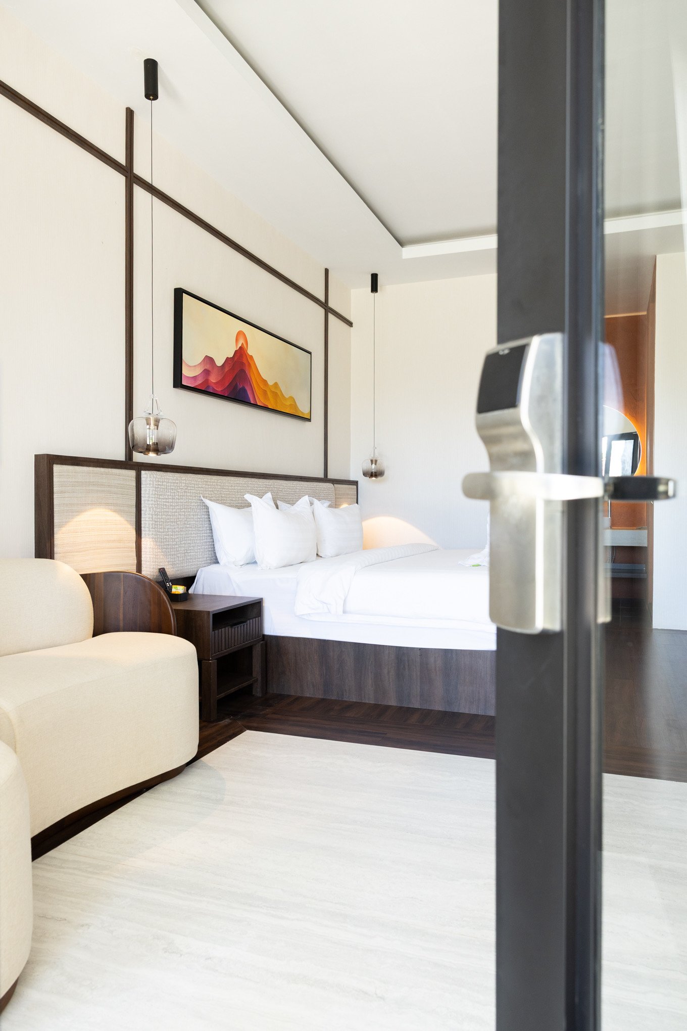 A hotel room with a bed, a small beige armchair, a nightstand with a telephone, a painting of a mountain landscape on the wall, and hanging pendant lights.