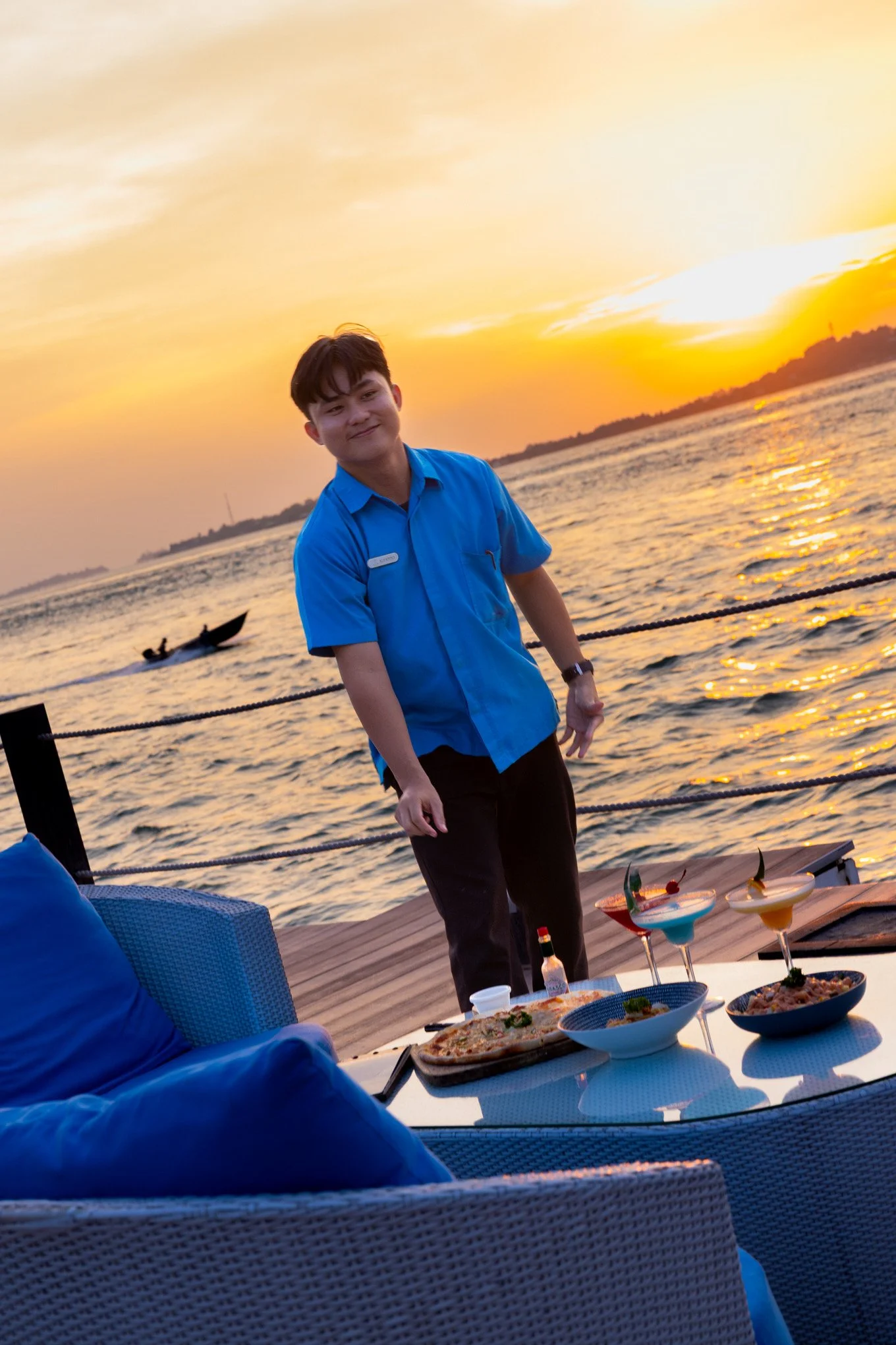 Young man in a blue shirt standing near a table with food and drinks, overlooking a sunset over a body of water.