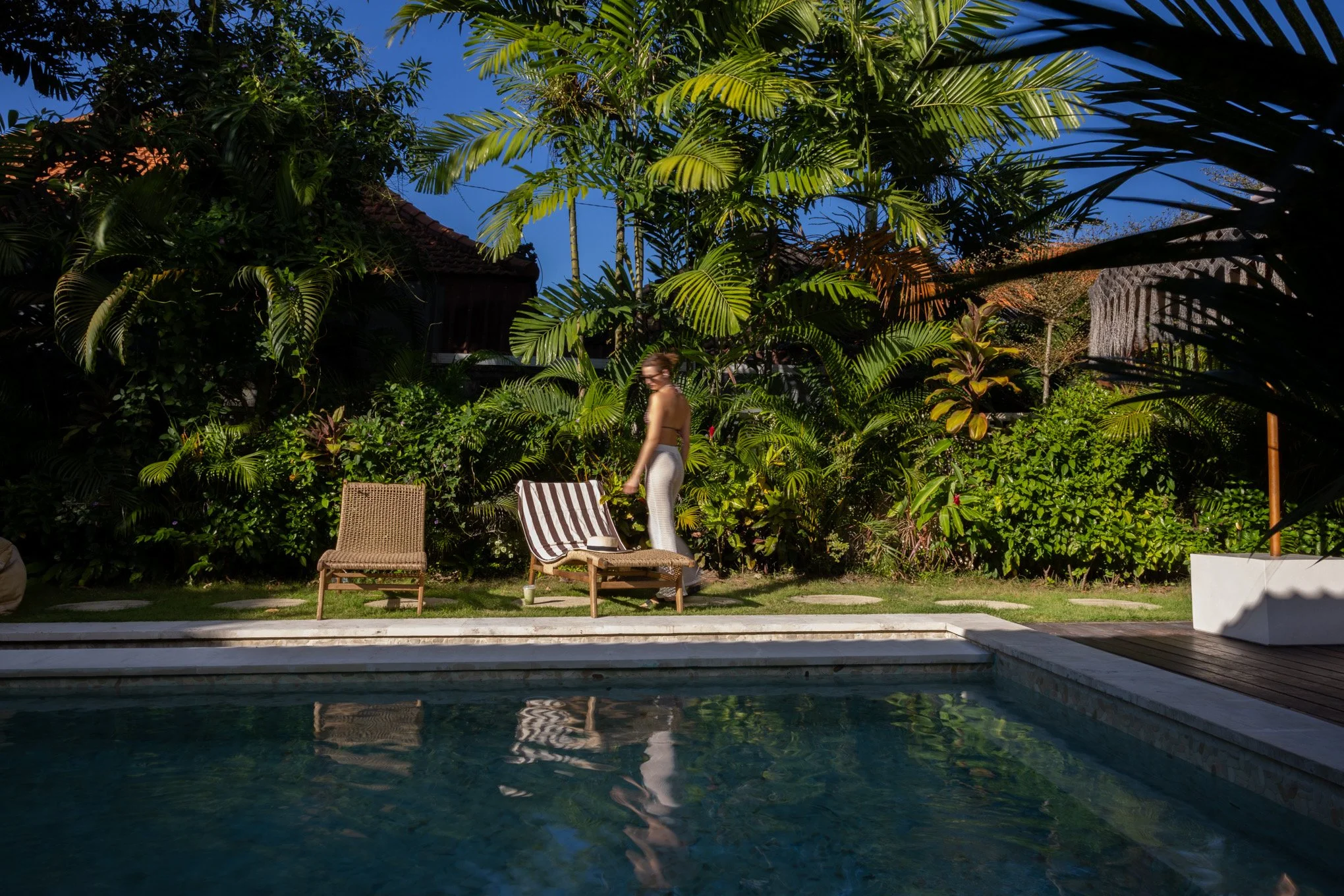 A woman in white and a bikini stands by a swimming pool in a lush backyard with dense tropical plants and trees against a clear blue sky.