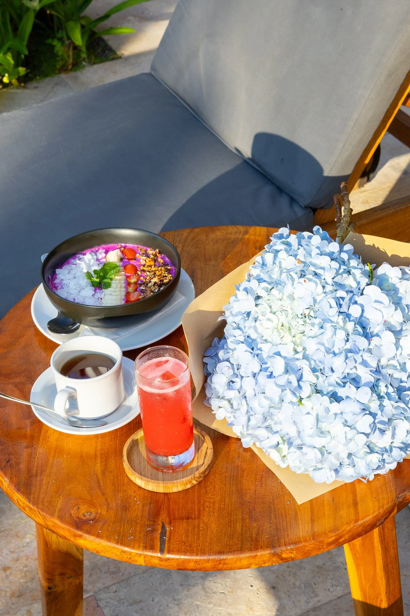 A wooden outdoor table with a bowl of yogurt topped with strawberries, bananas, granola, and a sprig of mint, a white coffee cup, a glass of pink juice, and a bouquet of white hydrangeas.