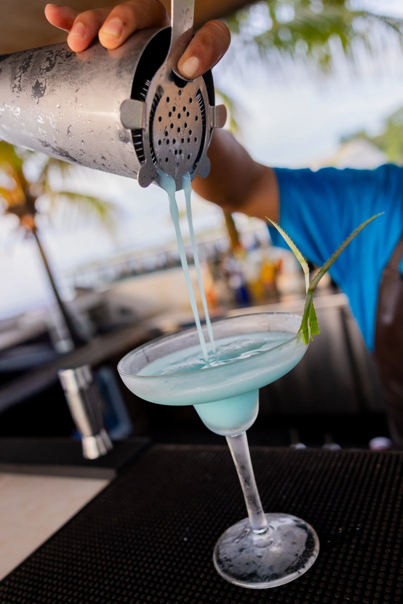 A person pouring a blue cocktail from a cocktail shaker into a martini glass garnished with a pineapple leaf, outdoors with palm trees in the background.