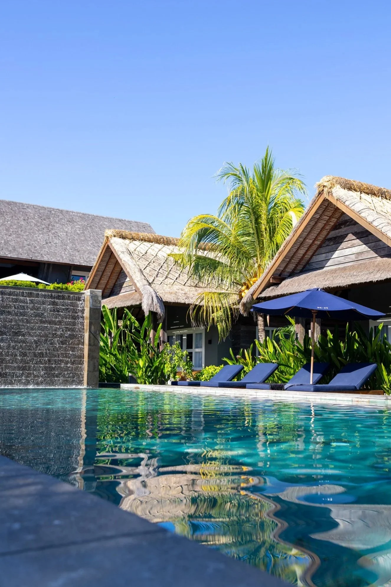 A resort-style swimming pool with blue lounge chairs and umbrellas, surrounded by tropical plants and thatched-roof cottages under a clear blue sky.