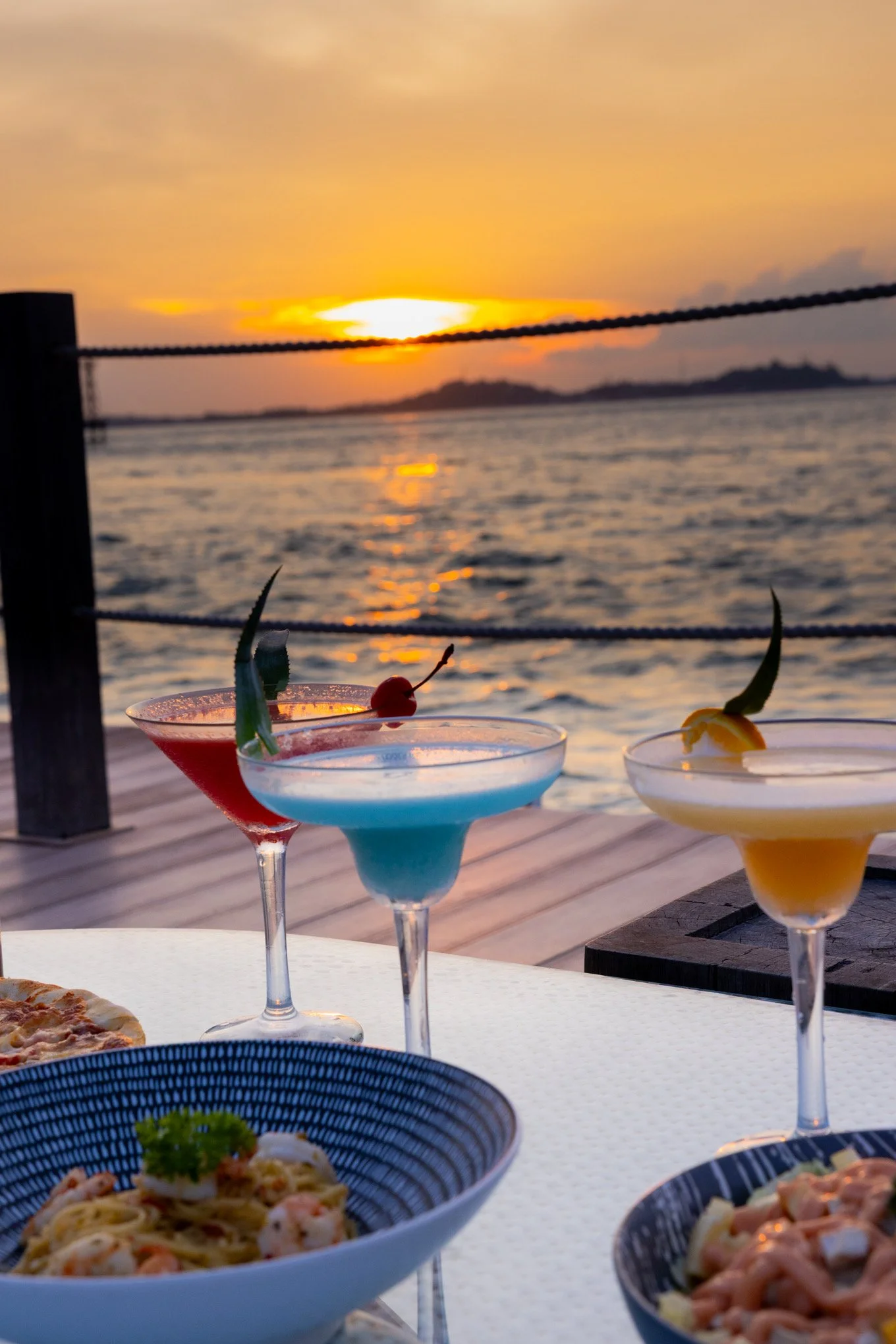Three colorful cocktails on a table near a body of water during sunset, with a blurred mountain in the background. There is a dish of pasta and a pizza in the foreground.