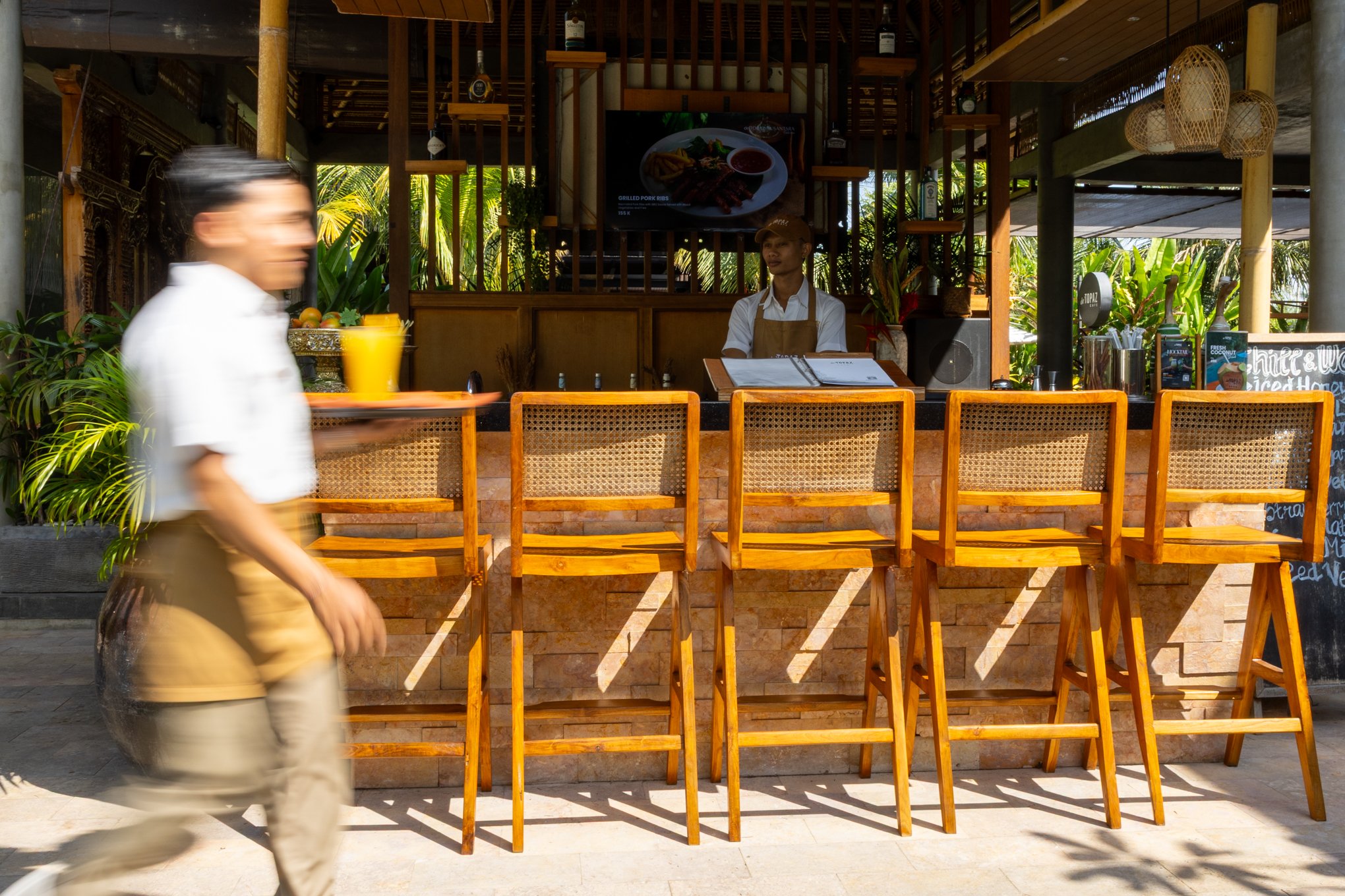 A blurred person walking past a wooden bar counter with empty chairs, a woman standing behind the bar, and tropical plants with bright sunlight in the background.