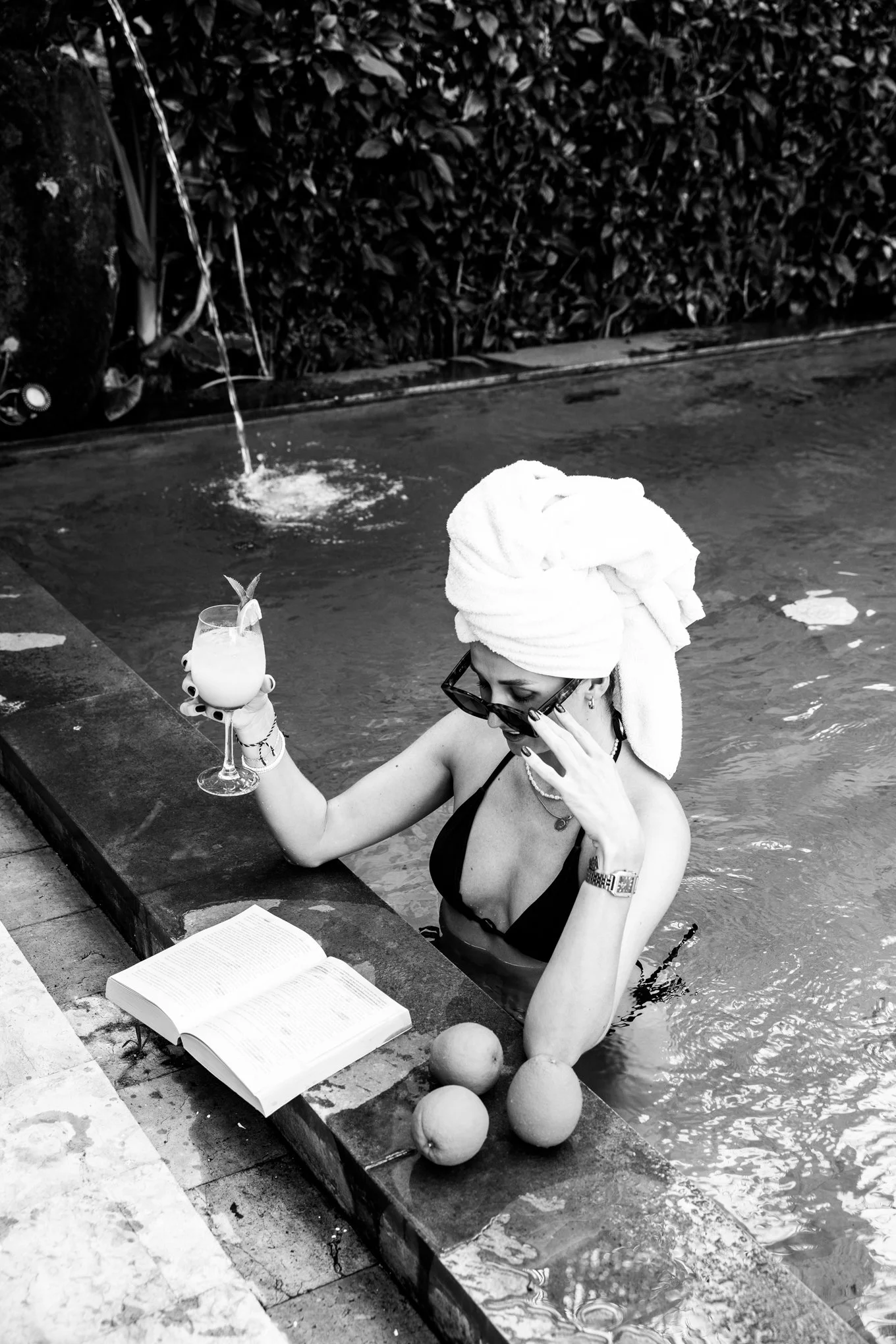 Woman with towel on head relaxing in a pool, reading a book, with a drink and fruits nearby.