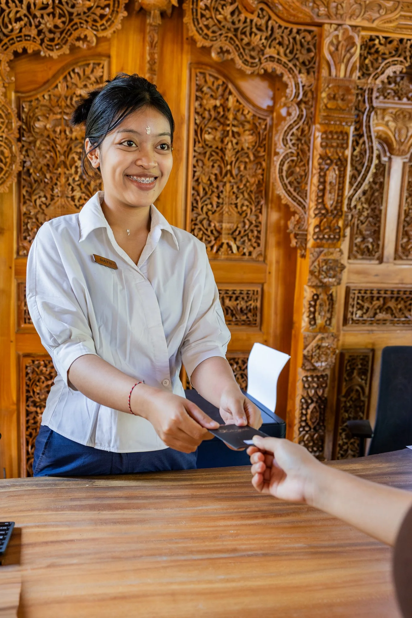 A woman with a name tag and braces is smiling and handing a receipt or card to someone across a wooden counter, with an ornate wooden wall in the background.