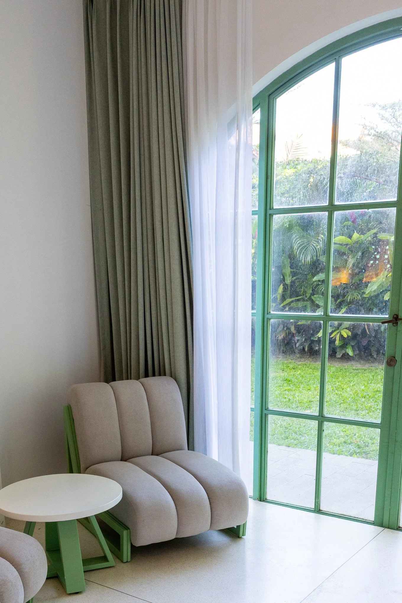 A cozy living room corner with a beige, plush armchair featuring green wooden accents, positioned next to a small round white side table. Behind the chair, there are elegant tall beige curtains and a sheer white curtain covering a large green-framed 