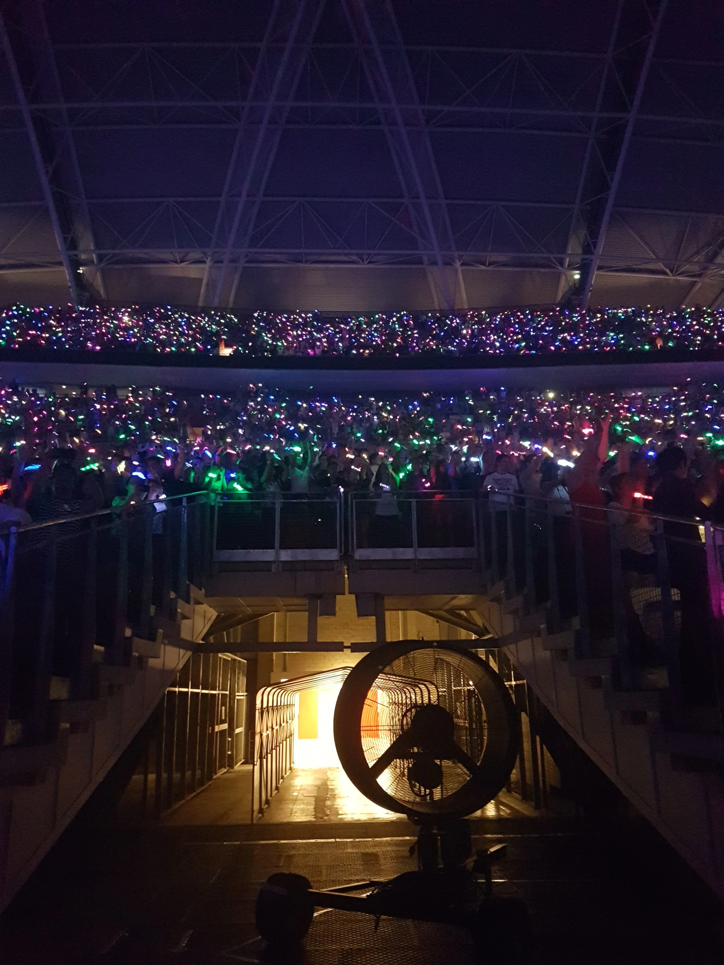 An audience holding LED lights at a stadium concert, seated over a tunnel with a large fan and a passageway illuminated by warm light at the end.