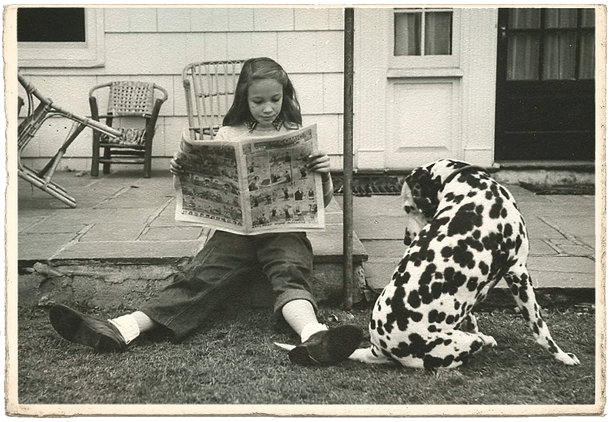 A young girl sitting on a patch of grass reading a comic book, with a Dalmatian dog sitting next to her, near a porch in a backyard.