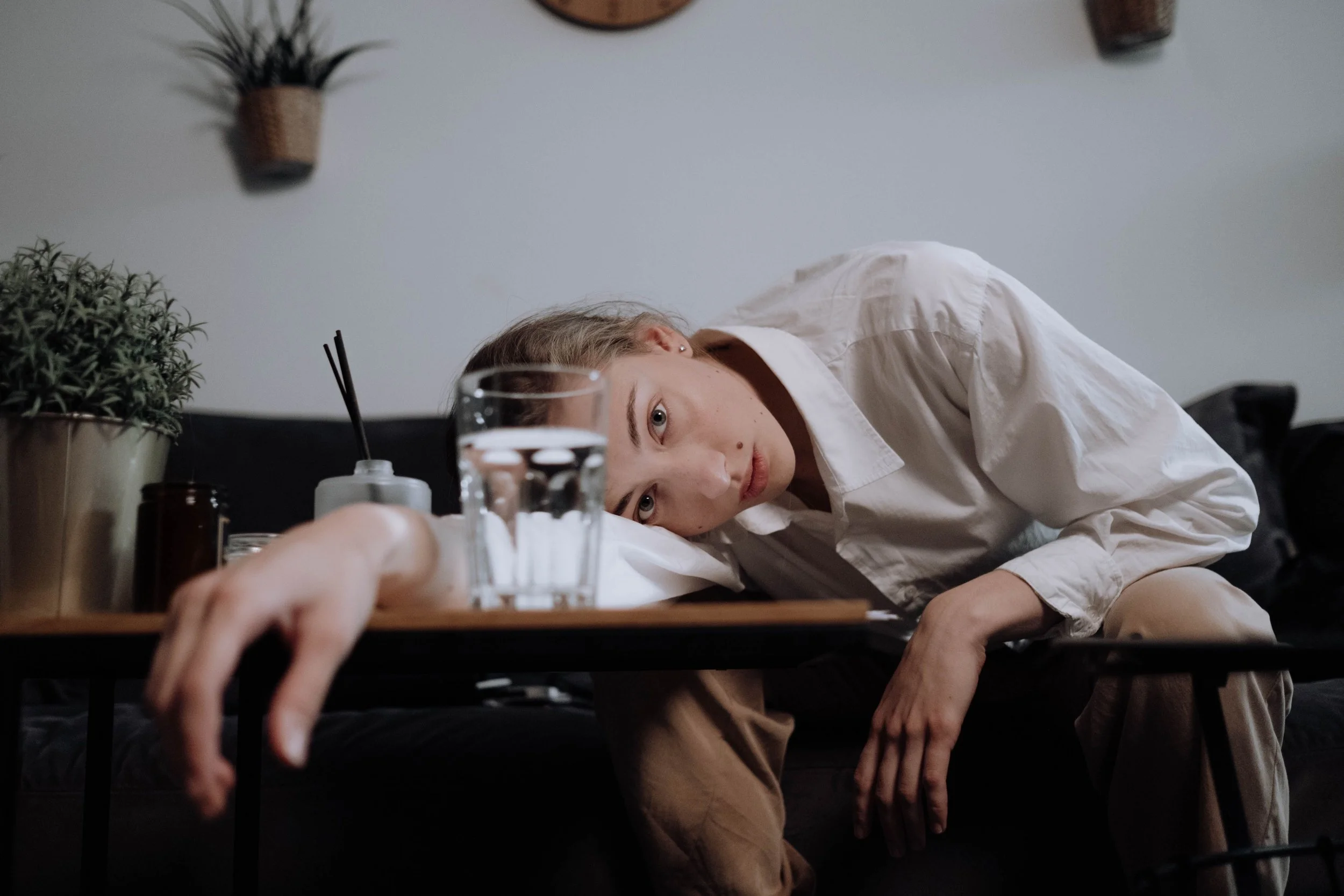 A woman with blonde hair and a white shirt lying across a dark table, reaching out with her arm, with a glass of water, a plant, and containers on the table. The background has a plain wall and some hanging baskets.