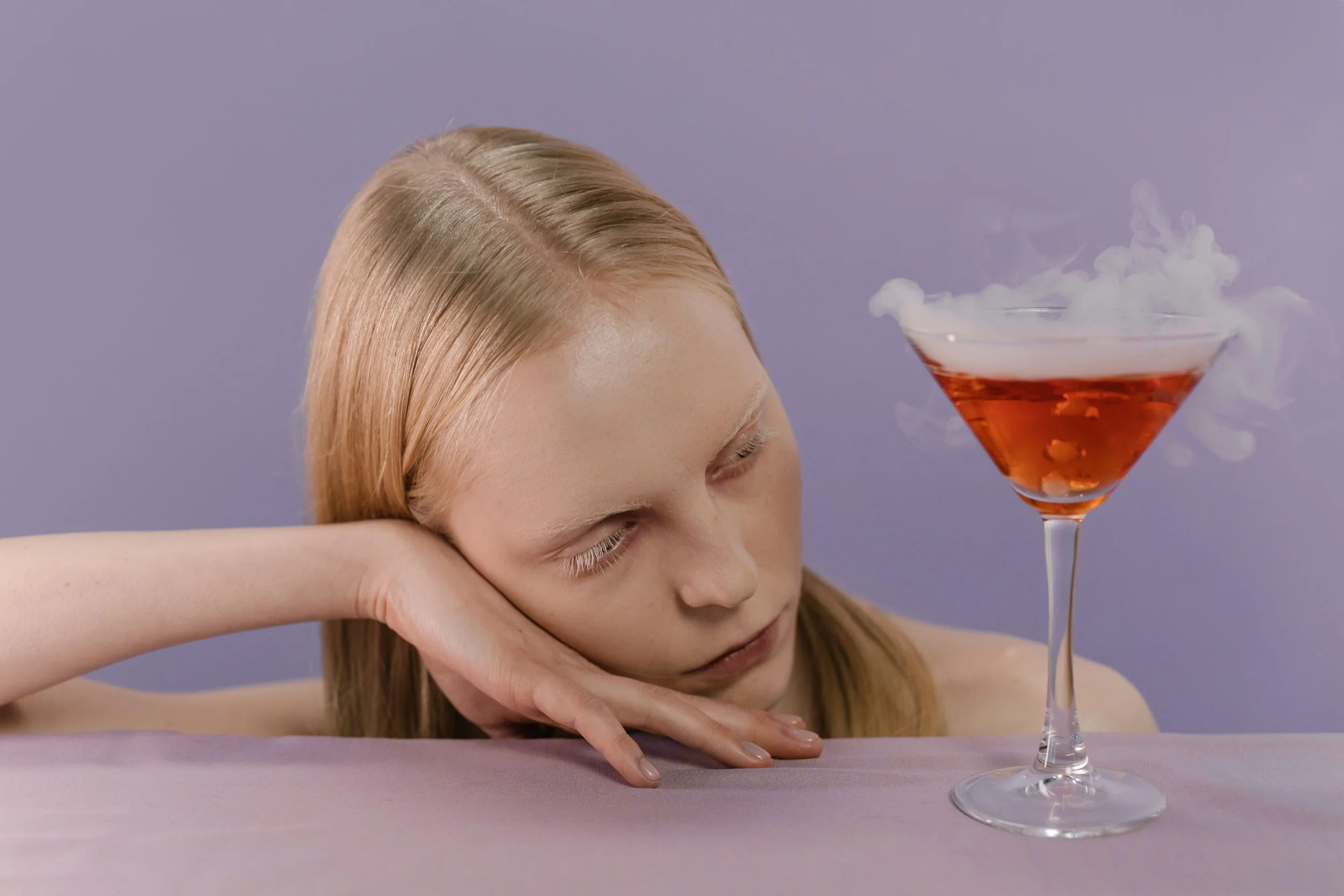 A woman rests her head on her arms with her eyes closed, next to a martini glass with a red cocktail and smoke coming from it, against a purple background.