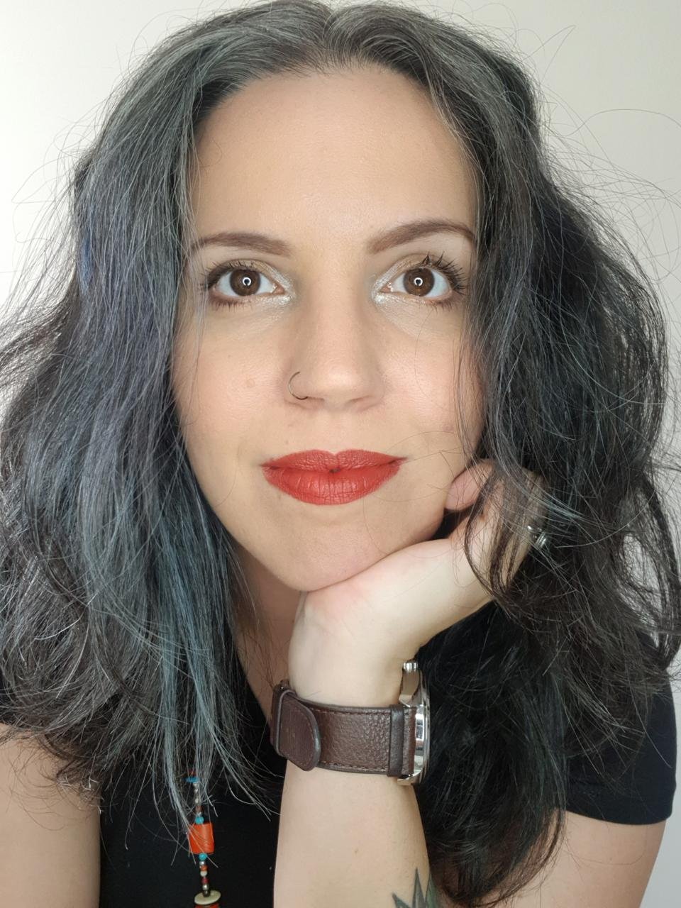 Close-up portrait of a woman with black and gray hair, wearing red lipstick, a nose ring, a brown wristwatch, and a beaded necklace, resting her chin on her hand.