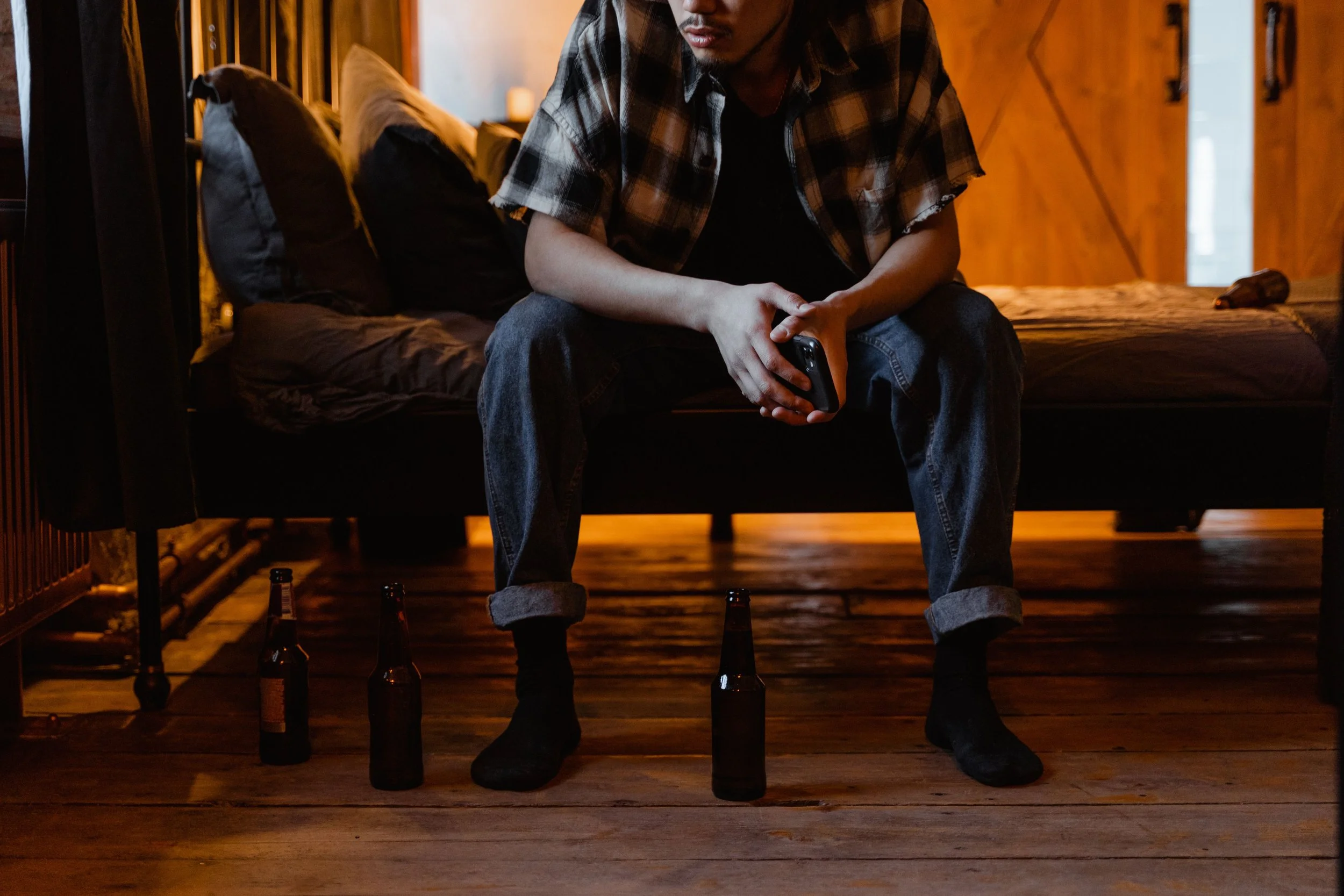 A man sitting on a bed holding a remote control in his hands, with beer bottles on the floor around him, in a dimly lit room with wood-paneled walls.
