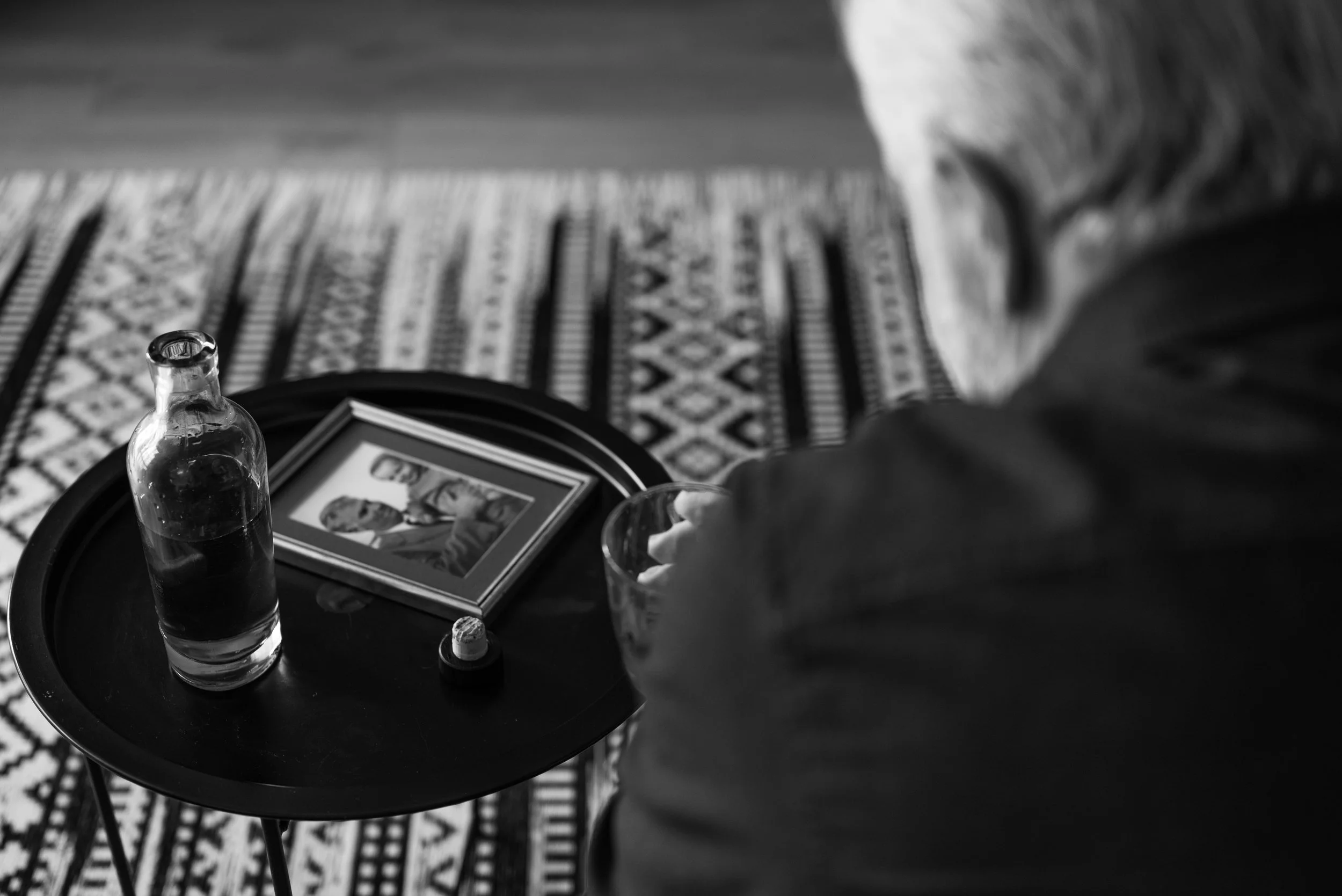 A black-and-white photo showing a table with a glass bottle, a framed photo, and a glass. In the background, there is a patterned rug on the floor and part of a person's arm and hand reaching toward the table.