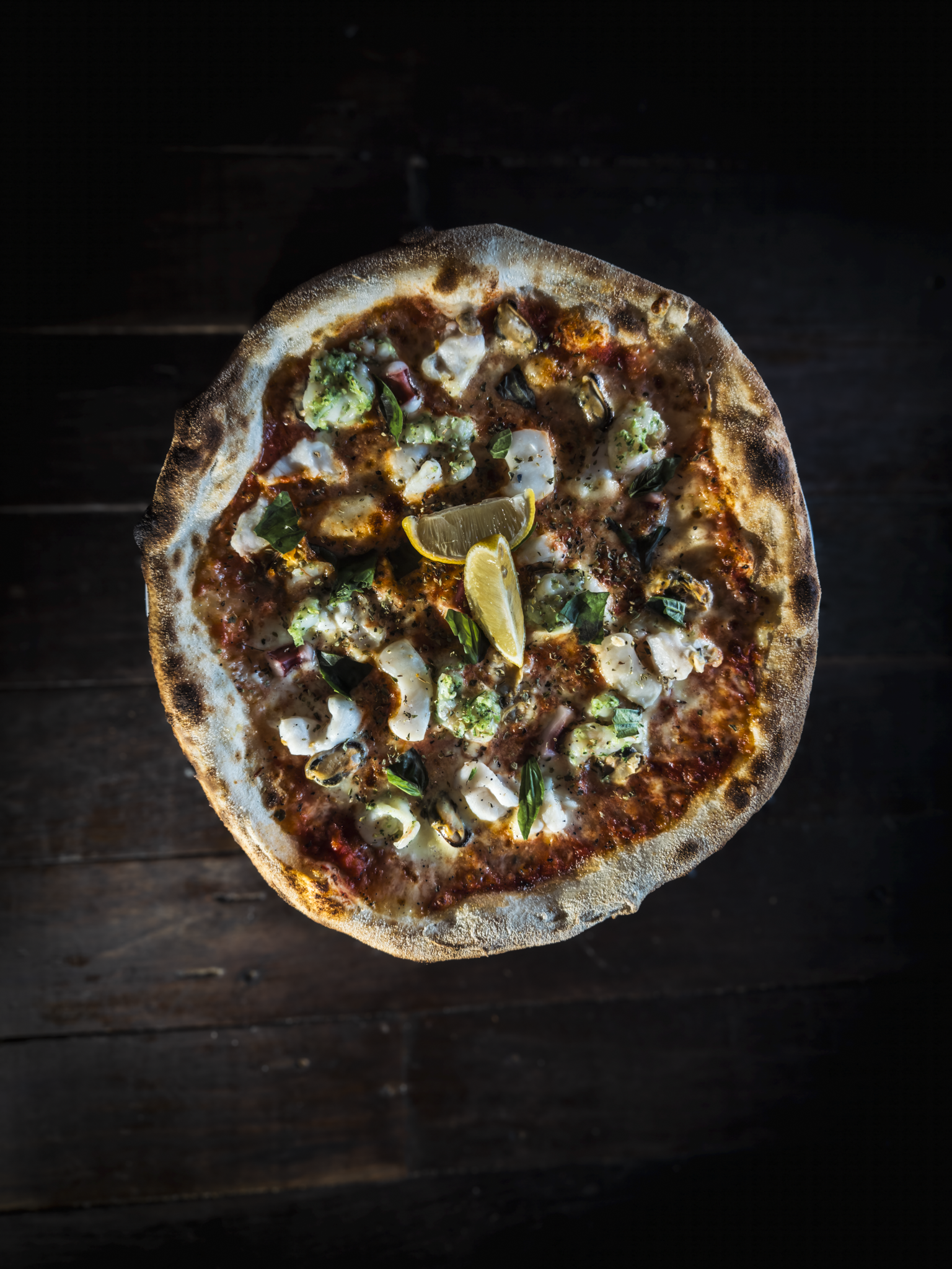 A pizza with various toppings such as broccoli, basil, cheese, and lemon slices on a dark wooden surface.